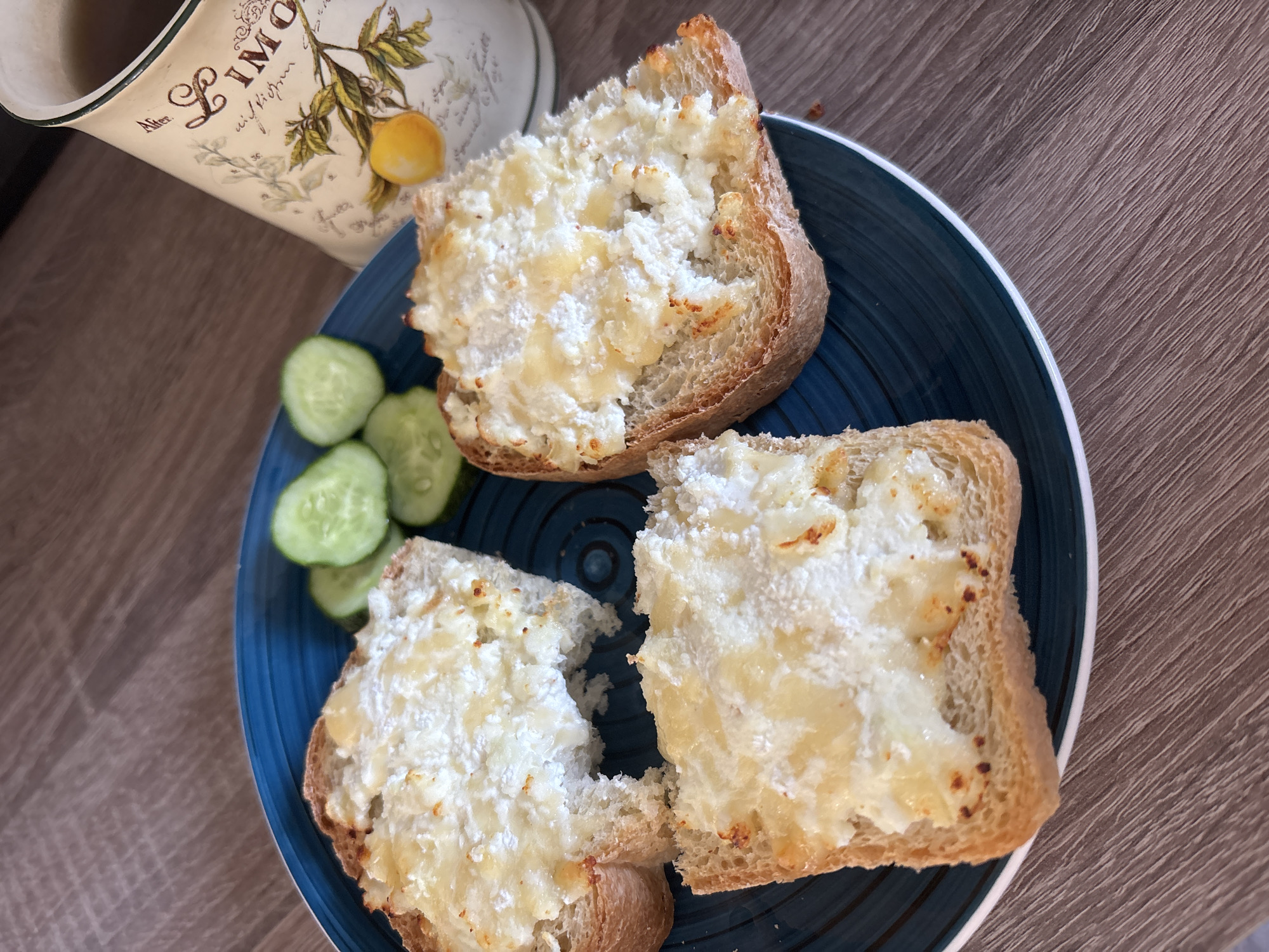 Three slices of toasted bread, topped with a white, creamy spread and a sprinkle of what appears to be melted cheese, are arranged on a blue, ridged plate. To the upper right of the plate are several slices of cucumber. A mug with a lemon-themed design and a dark liquid, likely tea, is situated behind the plate. The scene is set on a wooden surface. The image was taken in Naberezhnye Chelny, Russia.