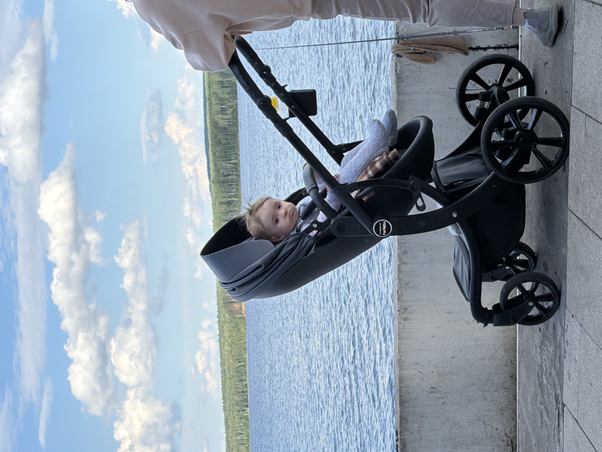 A baby sits in a black stroller by the Kama River in Chaikovsky, Russia. The baby, with light brown hair, wears a striped shirt and grey leggings. The stroller has a black frame and a deep grey bassinet. A person, only their lower body visible, stands behind the stroller, wearing a light beige hoodie and light grey pants. They appear to be holding a fishing rod. The background features a wide blue river with a distant shoreline of green trees under a sky with fluffy white clouds. A concrete barrier runs along the riverbank.