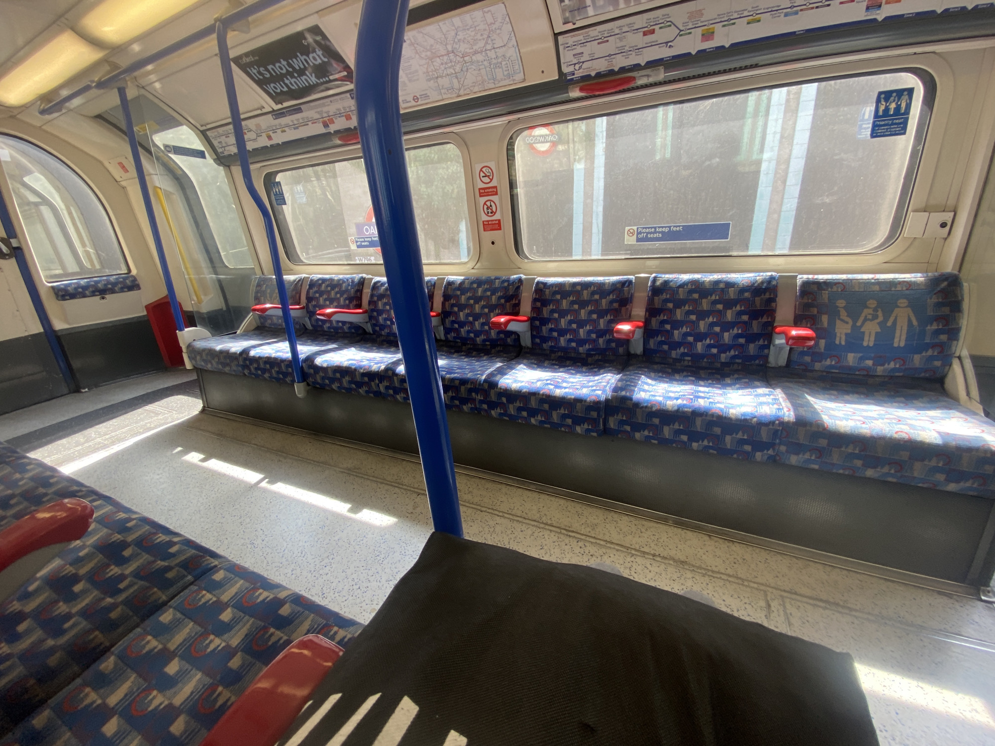 The image captures the interior of a London Underground train carriage, appearing mostly empty during a bright, sunny day in London, United Kingdom. Strong sunlight penetrates the windows, casting distinct shadows across the light-colored, speckled floor.
The main subjects are the train's furnishings: rows of characteristic blue and red patterned moquette seats with individual red grab handles. Blue vertical poles are positioned strategically throughout the carriage for standing passengers, although no people are visible in the frame.
Overhead panels above the windows display a visible London Underground map, indicating the train's context within the city's transport network. Various signs are attached to these panels and windows. Visible text includes an advertisement stating, "It's not what you think...", and multiple "Please keep feet off seats" notices. A "Priority seat" sign with pictograms for pregnant women, parents with young children, the elderly, and disabled individuals is prominent on a seatback. Other signs enforce "No smoking" and "No alcohol" policies via pictograms. A sticker with "W796" is visible near a window. The windows themselves show reflections and hints of the urban landscape outside, suggesting the train is either in motion or paused at a station. The overall impression is one of quiet transit, uncharacteristic of the usually bustling London Tube.