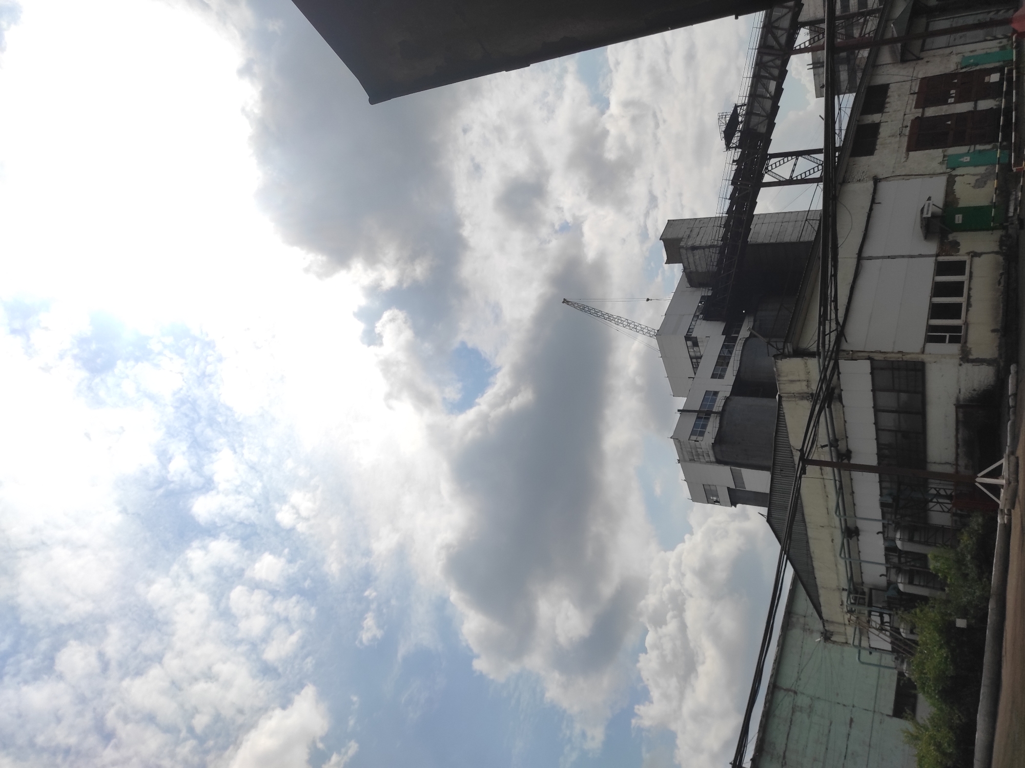 This is a low-angle, full shot of an industrial facility under a cloudy sky in Kyzyl, Kazakhstan. The foreground is dominated by the roofs and walls of industrial buildings. To the left, a light green, corrugated metal building with a slanted roof extends into the frame. In the center and right, a white and gray building complex with multiple stories and a large conveyor belt system is visible. A crane extends from the top of the central building. The sky is filled with a mix of white and gray clouds, with patches of blue sky visible. The scene appears to be a factory or processing plant, with evidence of ongoing industrial activity suggested by the crane and conveyor belt. No people are visible in the image.