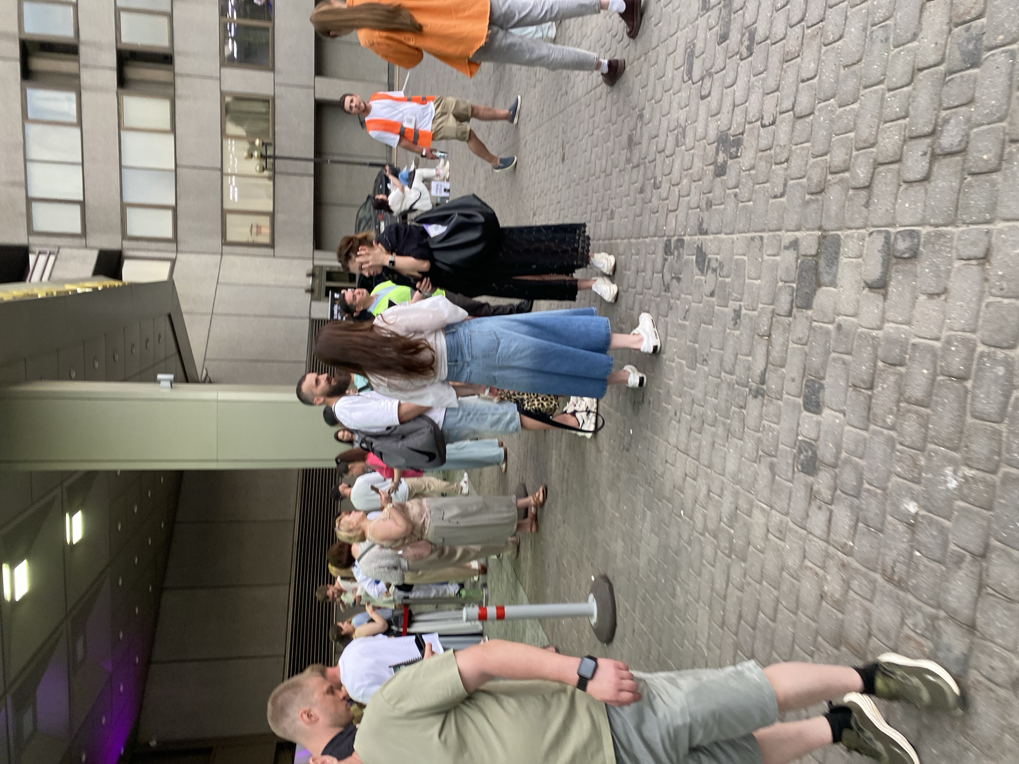 A medium-shot, outdoor image depicts a line of people on a cobblestone street in Moscow, Russia. The people are standing in an orderly fashion. Most face towards a building on the left, which has a modern architectural style, with many windowed floors. Some of the people are dressed in summer-wear clothing. There is a man at the front of the line wearing a light green shirt and shorts, and a black wrist watch. In the background, more people can be seen in the line. A man in an orange vest is toward the top of the frame. The sky is overcast and the building appears to be casting a shadow over the line.