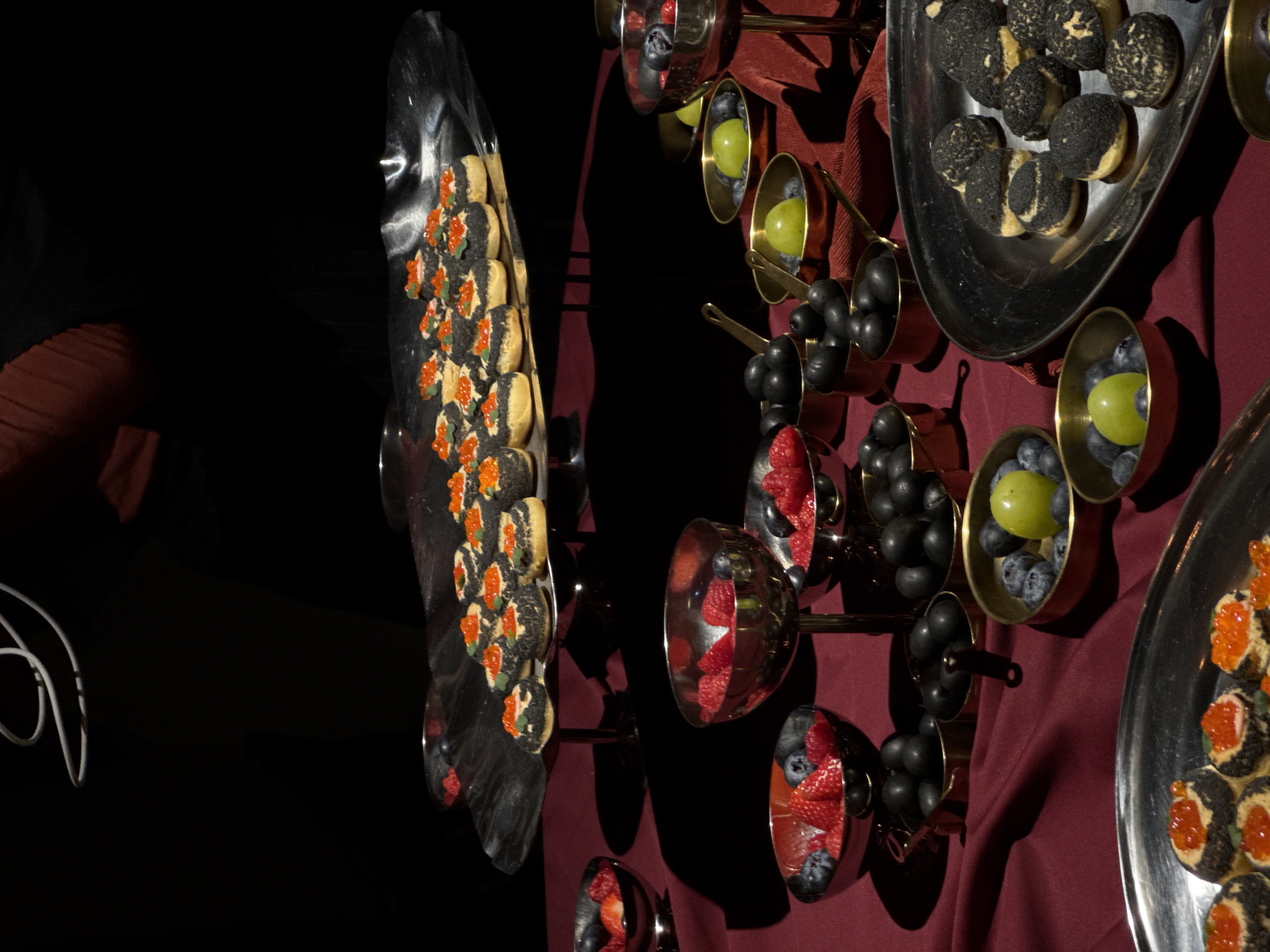 The image displays an indoor catering setup, likely at an event, in Yerevan, Armenia. A dark red tablecloth covers the display surface. The upper portion of a person, wearing a dark garment with a visible reddish-brown sleeve or lining, is partially visible at the top against a black background. A white, looped cable or object is suspended above the food display.
The primary focus is a large, reflective silver-colored tiered serving stand. It holds numerous small, circular canapés, each featuring a light-colored base, a dark-colored, textured coating, and topped with bright orange fish roe (caviar) and a tiny green garnish.
Arranged on the red tablecloth are various smaller serving vessels. Several reflective silver-colored bowls, some stemmed, contain a mix of fresh red raspberries and blueberries. Numerous small, brass-colored pots hold dark, spherical items, identified as black olives. Other brass pots contain a combination of blueberries and single bright green grapes or small green apples. A flat, elongated silver-colored tray to the right displays small, round pastries with a dark, cracked or textured topping. In the lower-left foreground, a portion of another silver-colored tray is visible, also holding canapés topped with orange caviar. The overall lighting is dim, suggesting an evening or atmospheric setting.