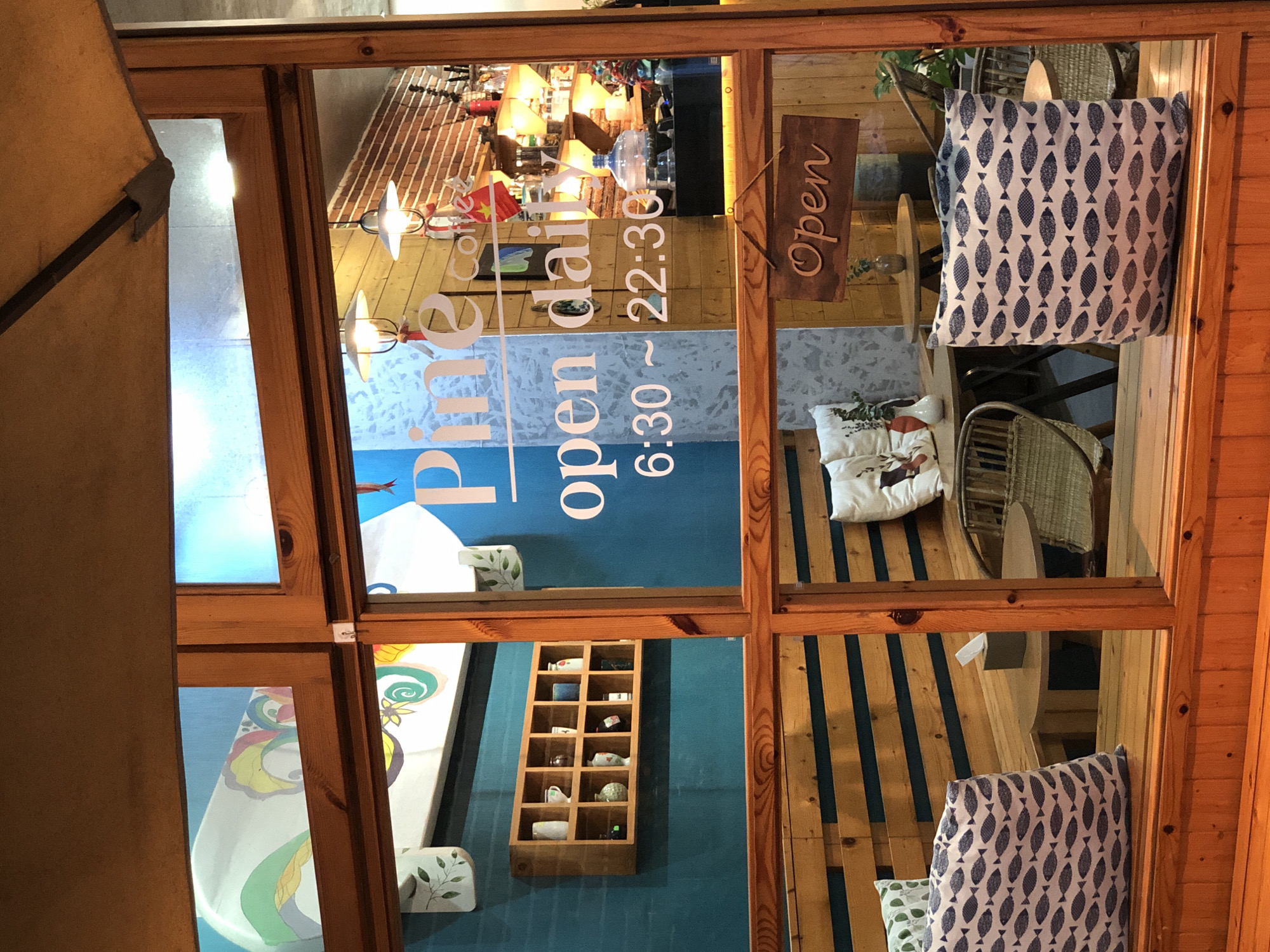 The image captures the exterior of a coffee shop with large glass windows, offering a view into its interior. The coffee shop appears to be located in Đà Nẵng, Vietnam, as suggested by the presence of a small Vietnamese flag visible behind the glass.
The window frames are made of warm-toned wood, and the glass displays white lettering indicating "Pine Coffee" and operating hours "6:30 ~ 22:30". A wooden sign that says "Open" in cursive is hanging from the frame. Inside, a brick wall is visible, along with shelves stocked with bottles and jars. Two pendant lights hang from the ceiling.
In the foreground, the view is partially obstructed by wooden benches and patterned cushions, one featuring a repetitive design of small, dark fish on a light background. There is also a tissue box on a small wooden table and a woven chair visible. The overall ambiance suggests a cozy and inviting atmosphere.