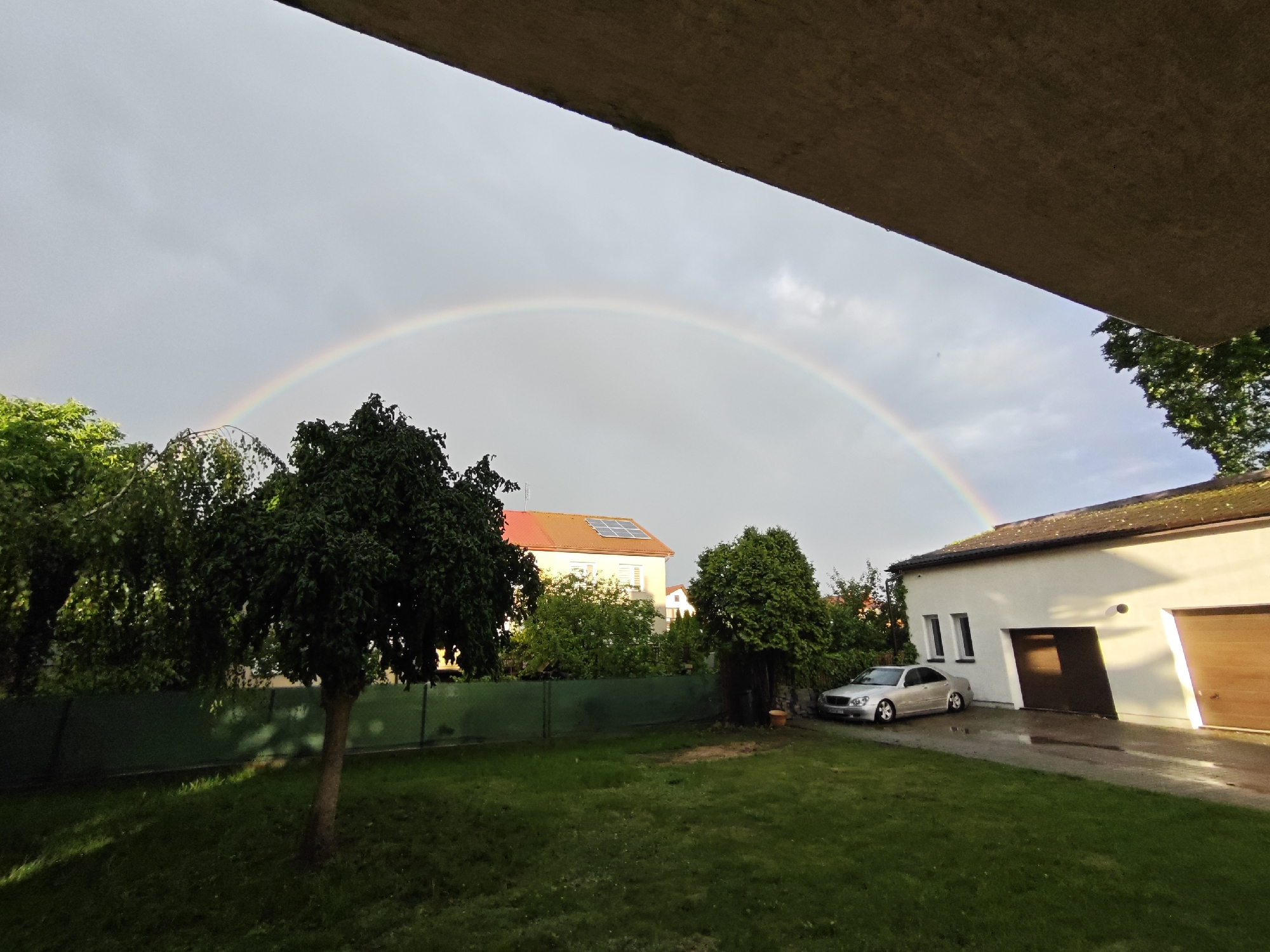 The image depicts a suburban backyard scene in Mława, Poland, under a cloudy sky with a visible rainbow arcing across the horizon. A silver sedan is parked in the driveway of a white building with a wooden garage door. The lawn is green and well-maintained, with several trees scattered throughout the yard. A dark green fence runs along the perimeter of the property. The sun appears to be setting, casting a warm glow on the building.