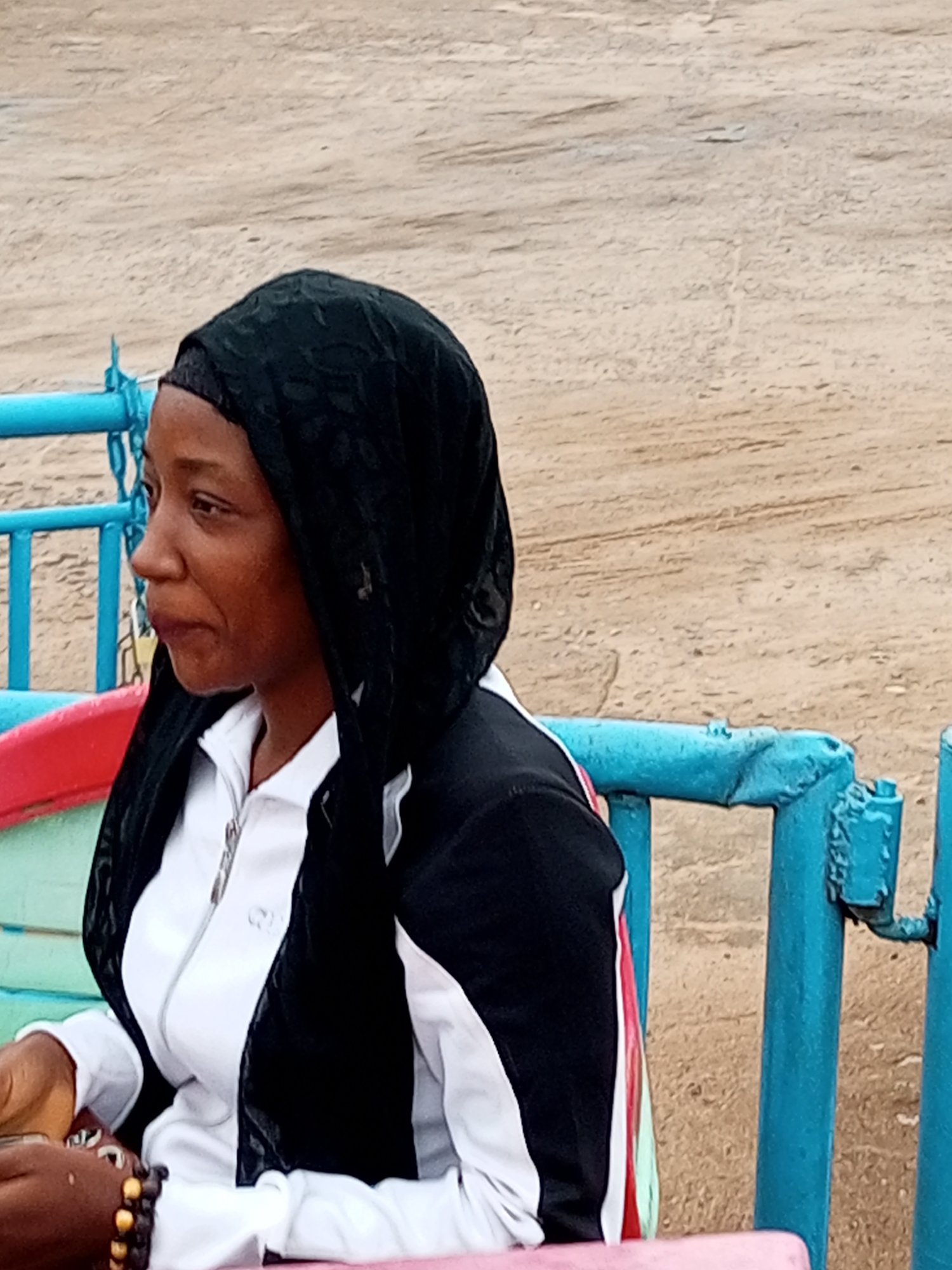 A young Nigerian woman, wearing a black headscarf and a black and white jacket over a white top, sits in a brightly colored vehicle with blue metal railings. Her left arm is bent at the elbow, and her left hand rests on the seat of the vehicle, adorned with a dark beaded bracelet. Her face is turned to the left, and she appears to be looking towards something outside the frame. The background is an expanse of dry, sandy ground, characteristic of the arid environment of Gombe, Nigeria. The vehicle she is in has a red and green trim on its side. The overall scene suggests an outdoor setting, possibly during a daytime outing in Gombe.