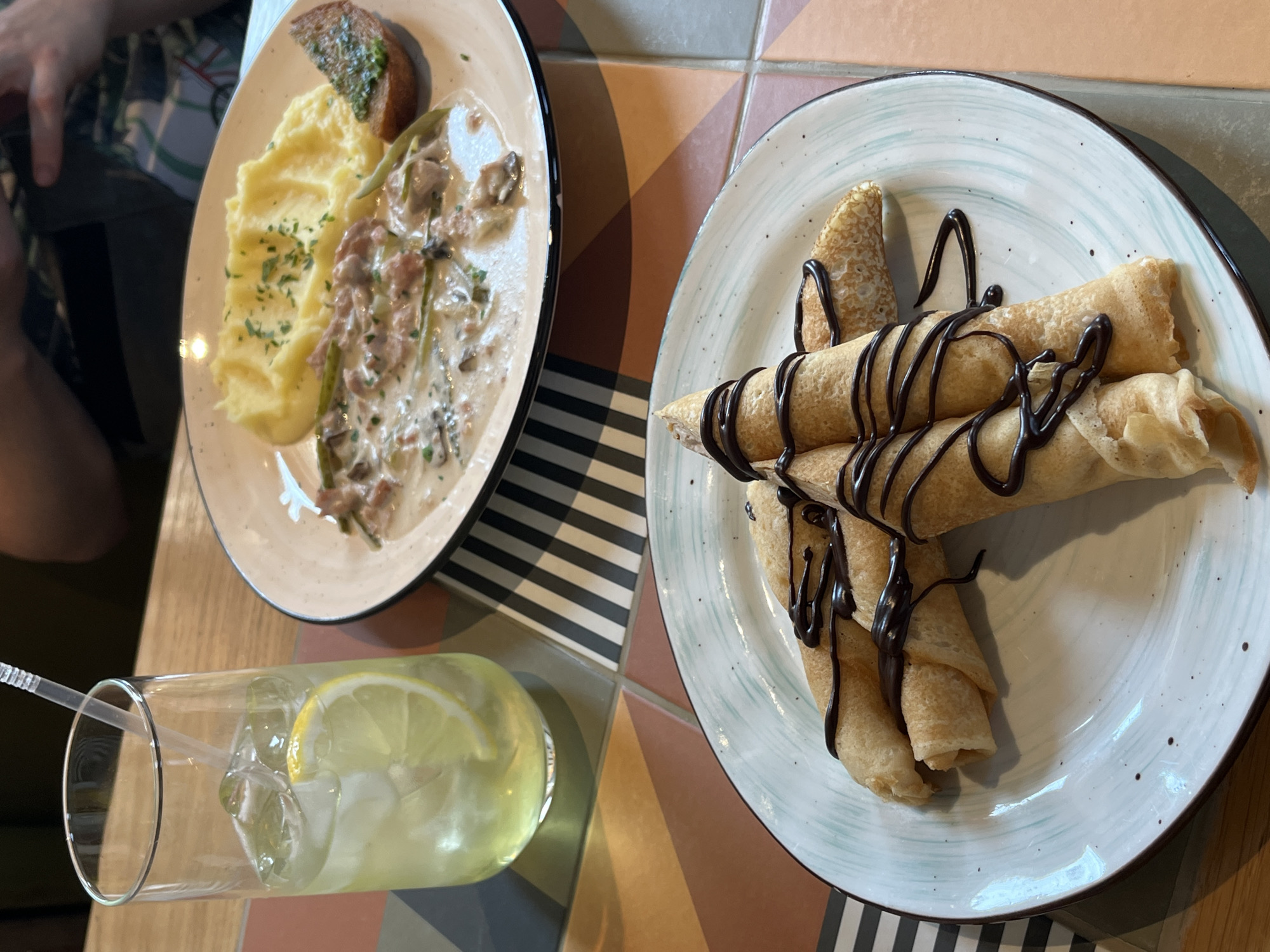 A high-angle shot shows two plates of food and a glass on a table. The plate on the left contains mashed potatoes with gravy, mushrooms, and greens, accompanied by a piece of toasted bread with herbs. The plate on the right holds several rolled crepes drizzled with chocolate sauce. Between the plates is a tall glass of lemonade with ice cubes and a lemon slice. The table surface has a patterned design of squares with alternating colors: black and white stripes, a brick-like orange-brown, and a light beige-brown. The image was taken in Verkhnyaya Pyshma, Russia.