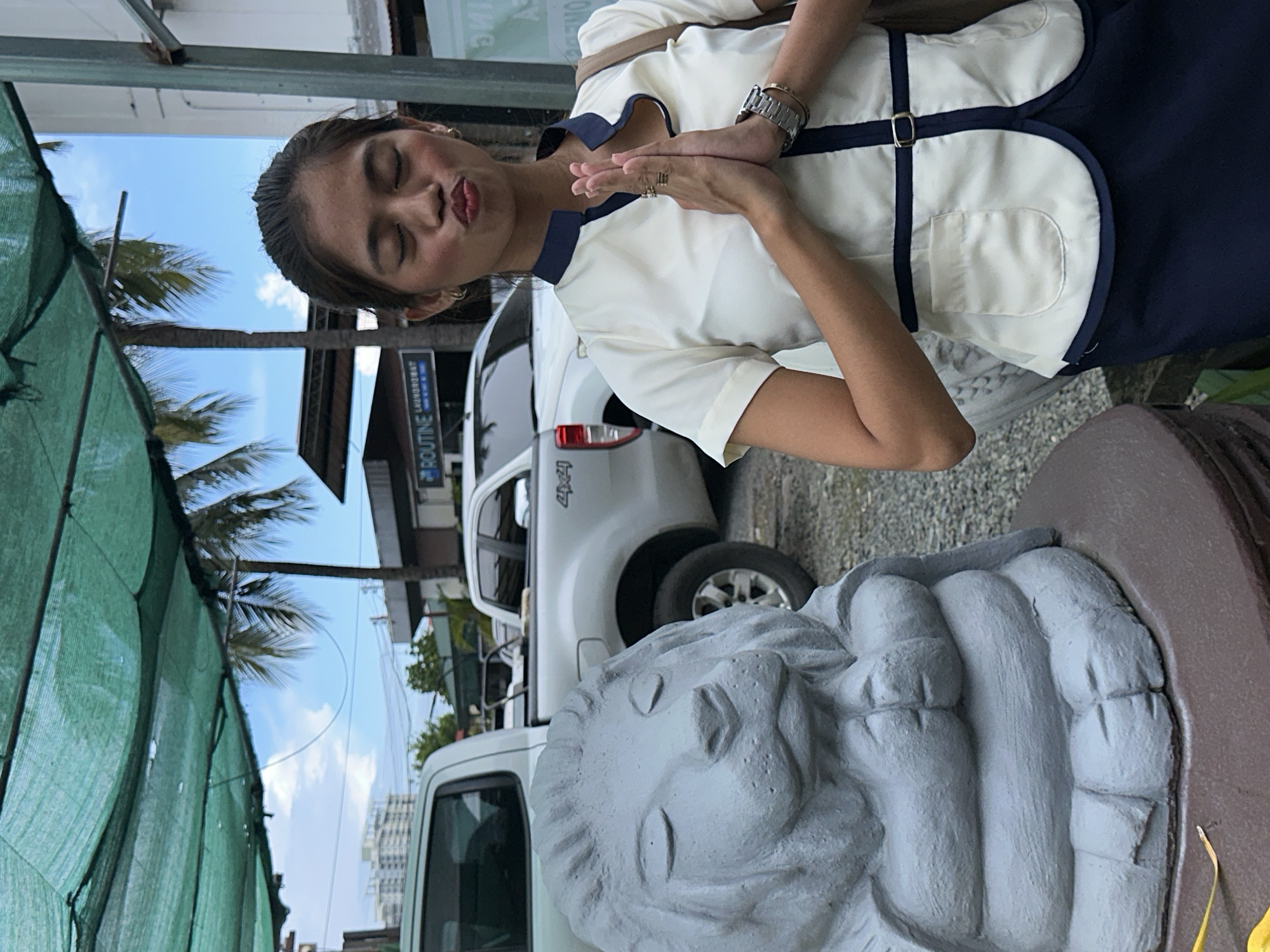A young woman in a white and navy uniform stands with her hands pressed together in a prayer-like gesture, puckering her lips, in front of a gray lion statue. The statue is intricately carved with layered folds resembling a mane and body. The woman is wearing a wristwatch and several rings. The scene appears to be outdoors, with a green mesh shade structure overhead, palm trees, and a blue sky with scattered clouds. In the background, two white pickup trucks are parked, and buildings are visible, including one with "ROUTINE LAUNDRY" signage. This setting is consistent with an outdoor commercial or public area in Davao City, Philippines, possibly near a business establishment or a park.