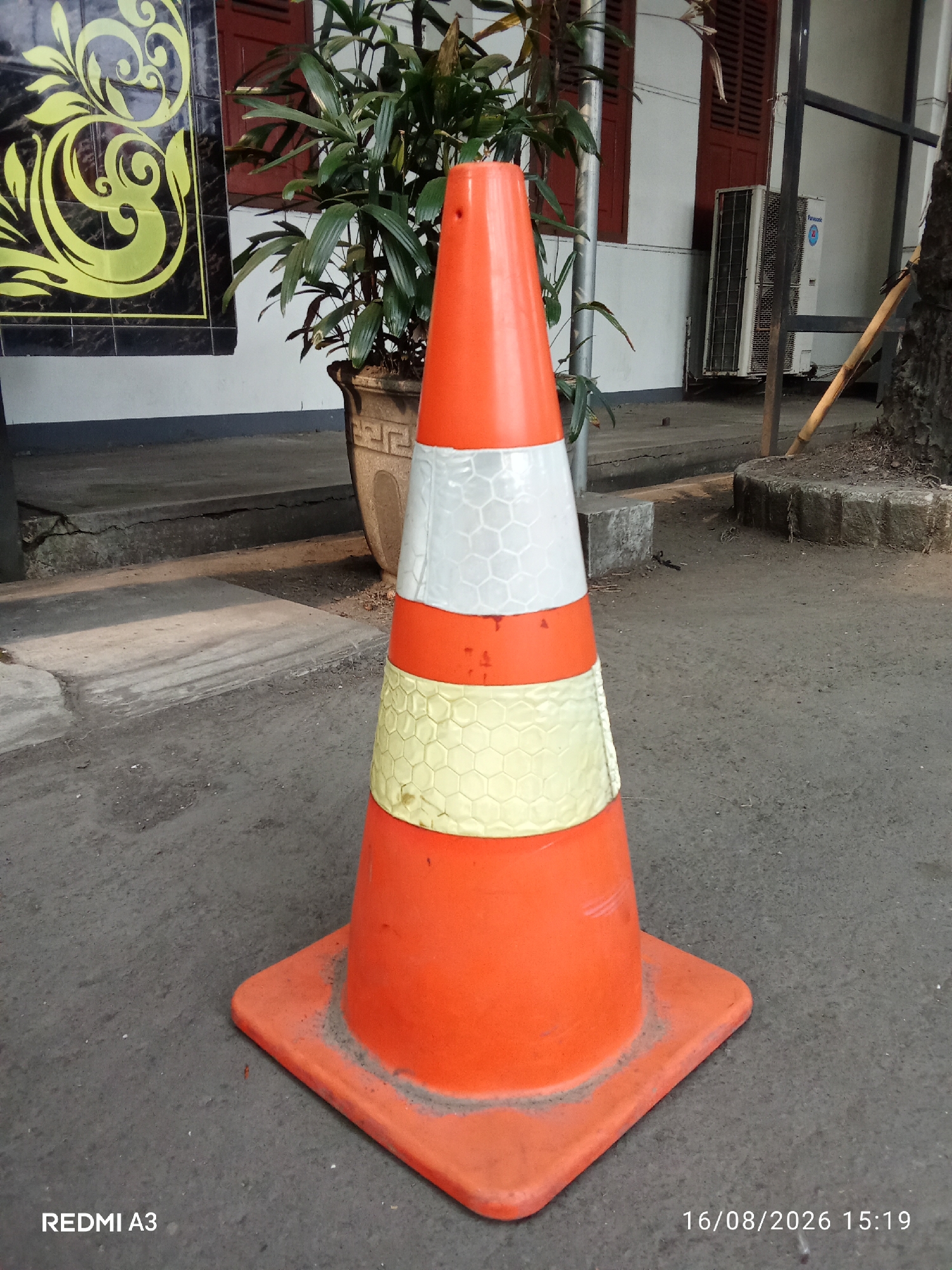 An orange traffic cone with two reflective bands, one white and one yellow, stands on asphalt pavement. The cone is square-based. In the background, a potted plant with green leaves is visible next to a wall with a black and yellow patterned decoration. An air conditioning unit is mounted on the wall further to the right. A wooden stick leans against a tree trunk to the far right. The timestamp "16/08/2026 15:19" and "REDMI A3" are present in the bottom corners of the image. The scene appears to be an outdoor urban environment in Surakarta, Indonesia.