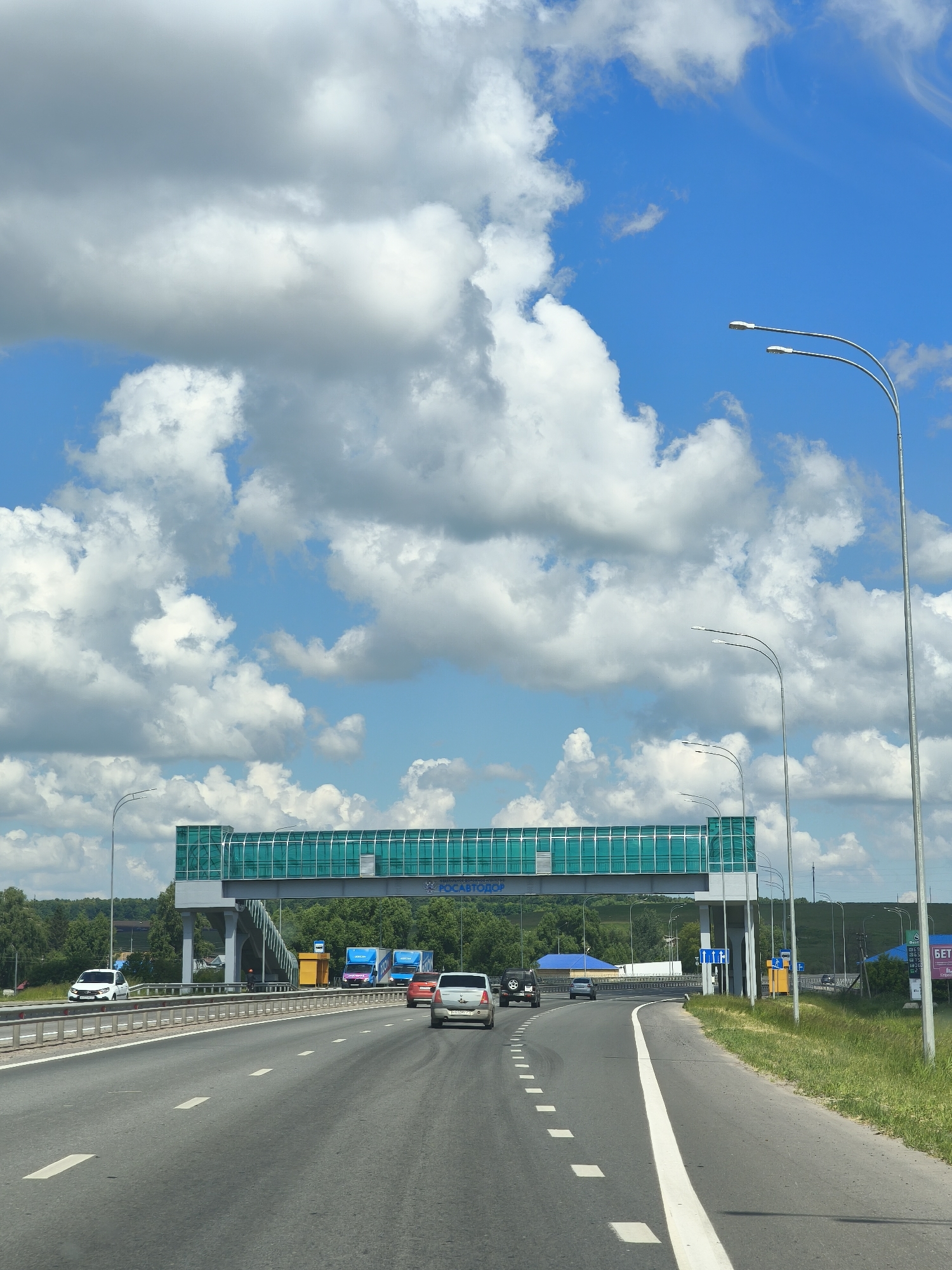 This is a medium-distance, eye-level, exterior, vertical shot taken from a vehicle on a highway in Shikhazany, Russia. The highway features multiple lanes and is bordered by a metal guardrail and a grassy shoulder. In the foreground, several cars are visible, driving in the same direction. A large pedestrian bridge with green tinted glass spans the highway in the middle ground. Beyond the bridge, a treeline and rolling hills are visible under a bright blue sky filled with large, puffy white clouds. Streetlights and road signs are also present.