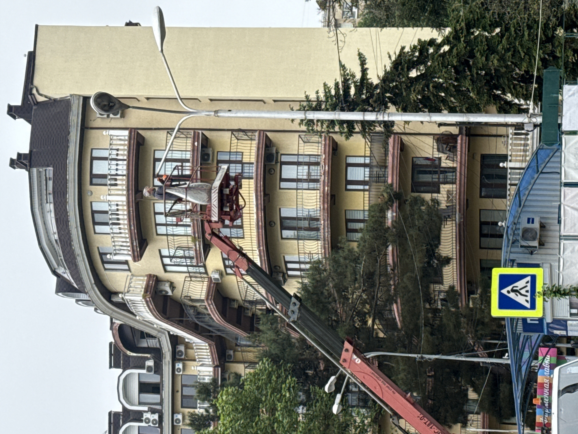 The image depicts a street scene in Unknown, Unknown, featuring a curved, multi-story building painted in shades of yellow and beige, with dark brown accents. The building has numerous windows and balconies, and air conditioning units are visible on the exterior.
A red crane is positioned in front of the building, with a worker in a lift basket near the upper floors. A street lamp extends along the side of the building. Green trees partially obscure the building, adding a natural element to the urban landscape.
In the foreground, a pedestrian crossing sign is prominently displayed. Parts of a building are seen in the right side of the frame. The overall atmosphere is somewhat overcast and indistinct due to the gray sky.