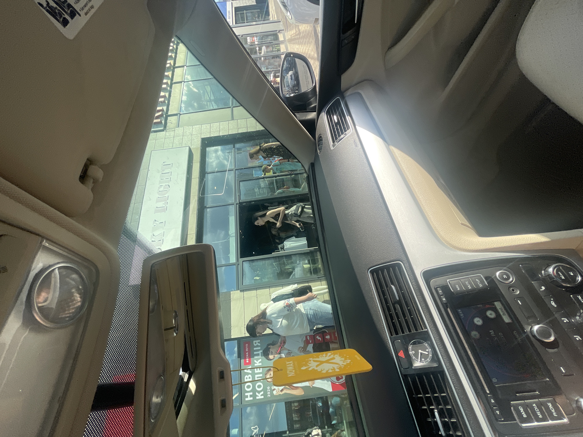 This media file captures an interior view of a car, likely parked in Kyiv, Ukraine. The scene is viewed through the windshield and side windows, reflecting the external environment.
The primary subjects visible are several people whose reflections are seen in the large glass facade of a building. These individuals appear to be indoors, possibly shopping or working, with one person in the foreground wearing a white shirt with embroidered details and jeans, looking down. Another reflection shows a person in a darker outfit.
Several objects are also identifiable. Inside the car, the dashboard and a portion of the steering wheel are visible, with a prominent infotainment screen displaying radio information and buttons for navigation, media, and phone functions. A small analog clock is affixed to an air vent. A yellow car air freshener with the text "NOWAX" is hanging from the rearview mirror.
The external setting is a modern building with large glass windows, suggesting a commercial or retail space. Text visible on the building's facade includes "KY LIGHT", "NOWAX", and what appears to be "НОВА ВЕСНА-ЛІТО КОЛЕКЦІЯ 2026", indicating a "New Spring-Summer Collection 2026". The reflection in the side mirror shows cars and the building's exterior, hinting at a bright, daytime scene. The overall impression is of a typical urban environment on a clear day.