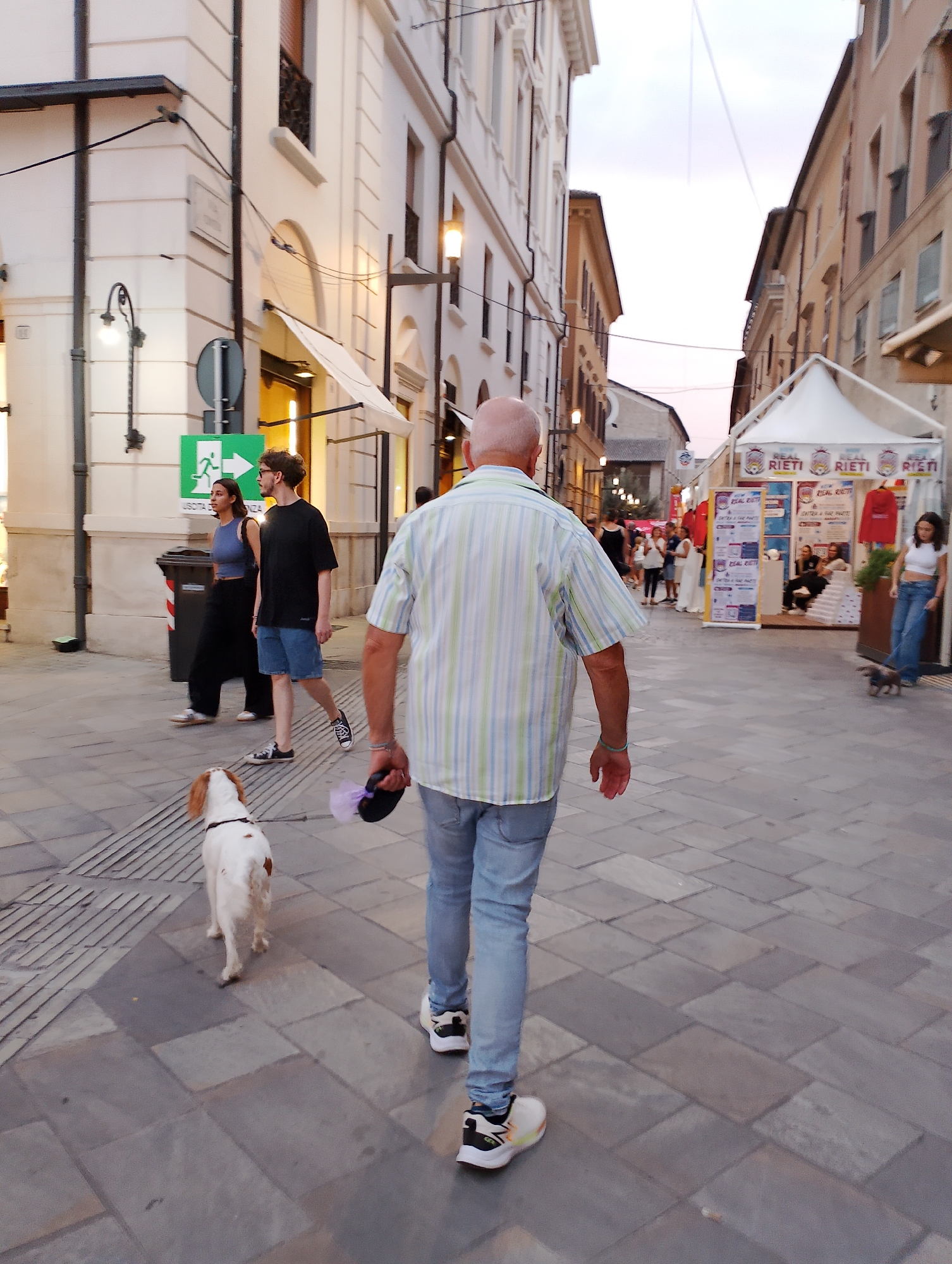 A man walks away from the camera down a city street in Rieti, Italy, his white and blue striped shirt and light blue jeans visible. He holds a purple leash in his right hand, attached to a small white dog with brown spots walking ahead of him. In the background, other pedestrians are visible, including a couple walking together, and a young woman with a small dog on a leash. Tents and stalls are set up along the street, some displaying the "RIETI" logo. The architecture suggests an Italian urban setting with buildings lining both sides of the street. The sky is overcast with a pale hue.