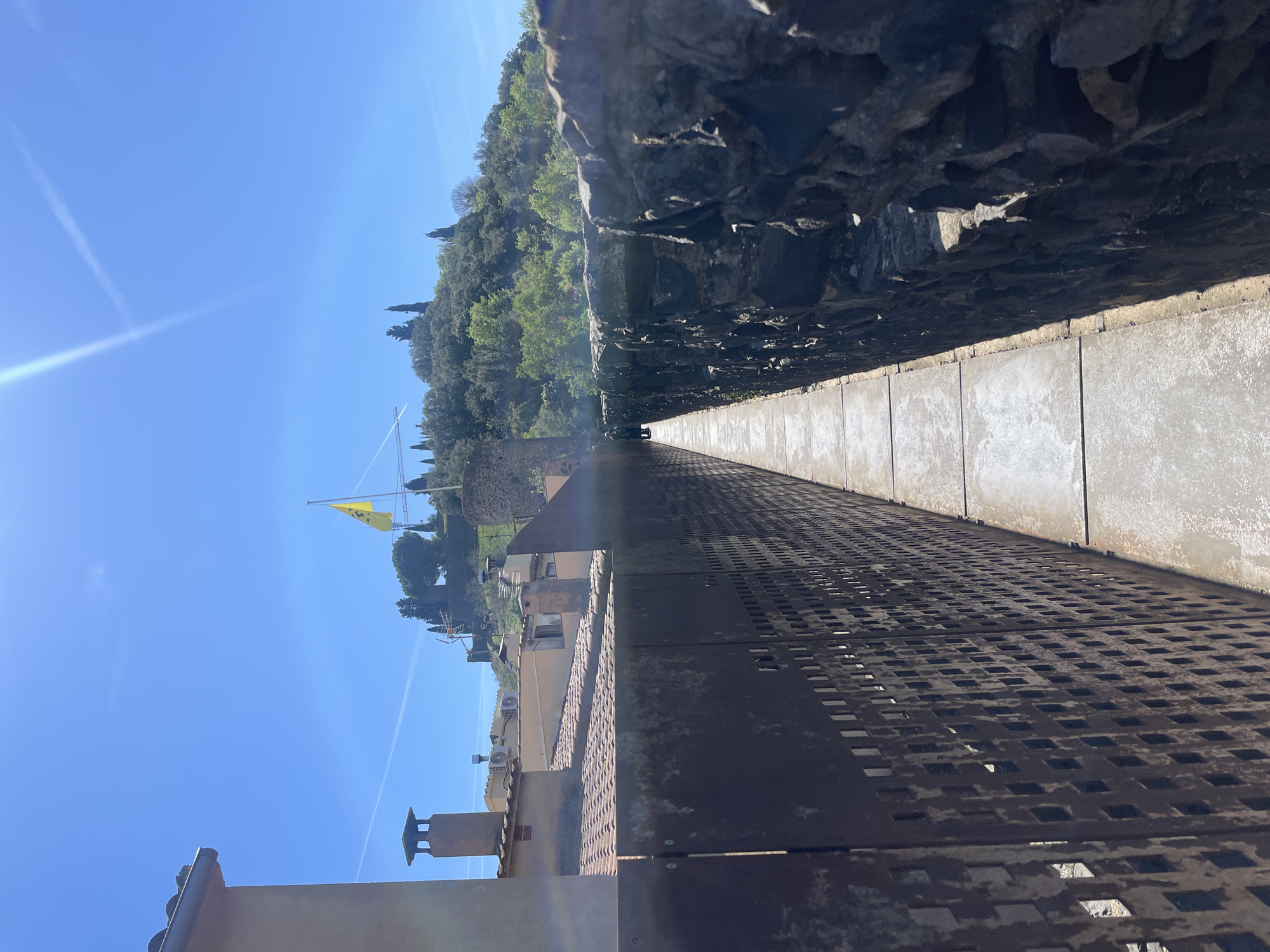 The image shows a view from the top of the medieval wall in Hostalric, Spain. The perspective is along the length of the fortified wall, with a walkway made of rectangular slabs running between the rough stone inner wall and a metal grate parapet on the outer edge. Beyond the wall, there are tiled rooftops and other buildings. The sky is clear blue. A yellow flag is visible on a tower in the distance. Trees and vegetation cover the landscape beyond the town.