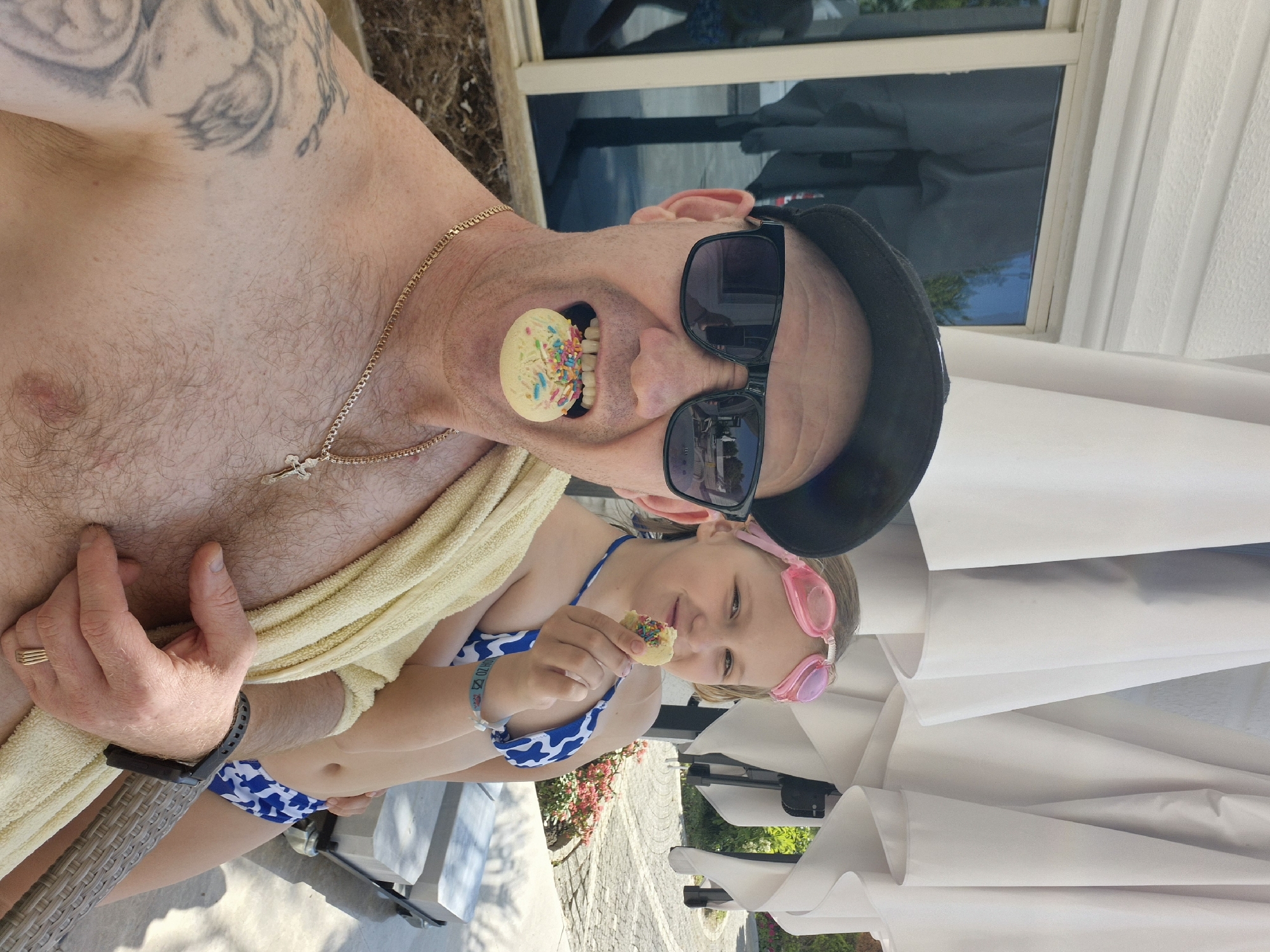 An outdoor scene in what may be Alanya, Turkey, shows a shirtless, light-skinned man wearing dark sunglasses and a black cap, holding a cookie in his mouth. He also has a gold chain around his neck and a towel draped over his chest. To his side, a young girl in a blue and white swimsuit and pink goggles holds a similar cookie. The setting includes a patio with outdoor furniture, flowers, a window and an umbrella.