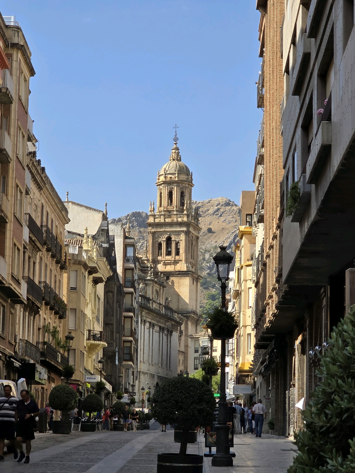 This is an eye-level, medium shot capturing a street scene in the city of Jaén, Spain. The perspective is looking down a pedestrianized street lined with multi-story buildings on both sides. The architecture is a mix of styles, with some buildings appearing older with decorative facades and balconies, while others are more modern and utilitarian.
Dominating the background is a large, ornate tower, likely part of a cathedral or church, with a distinctive domed top. Behind the tower, a rugged, sun-drenched mountain range forms the backdrop. The sky is a clear, bright blue with no clouds visible.
In the foreground and midground, people are strolling along the wide, paved street. Several individuals are walking in pairs or small groups, some appearing to be tourists. There are also potted trees and lampposts lining the walkway, adding to the urban ambiance. Some of the ground-floor buildings appear to be shops or cafes, with awnings and signs visible.
The lighting suggests it is daytime, likely late morning or early afternoon, with sunlight casting shadows and highlighting the architectural details. The overall impression is of a vibrant, historic city center on a sunny day.