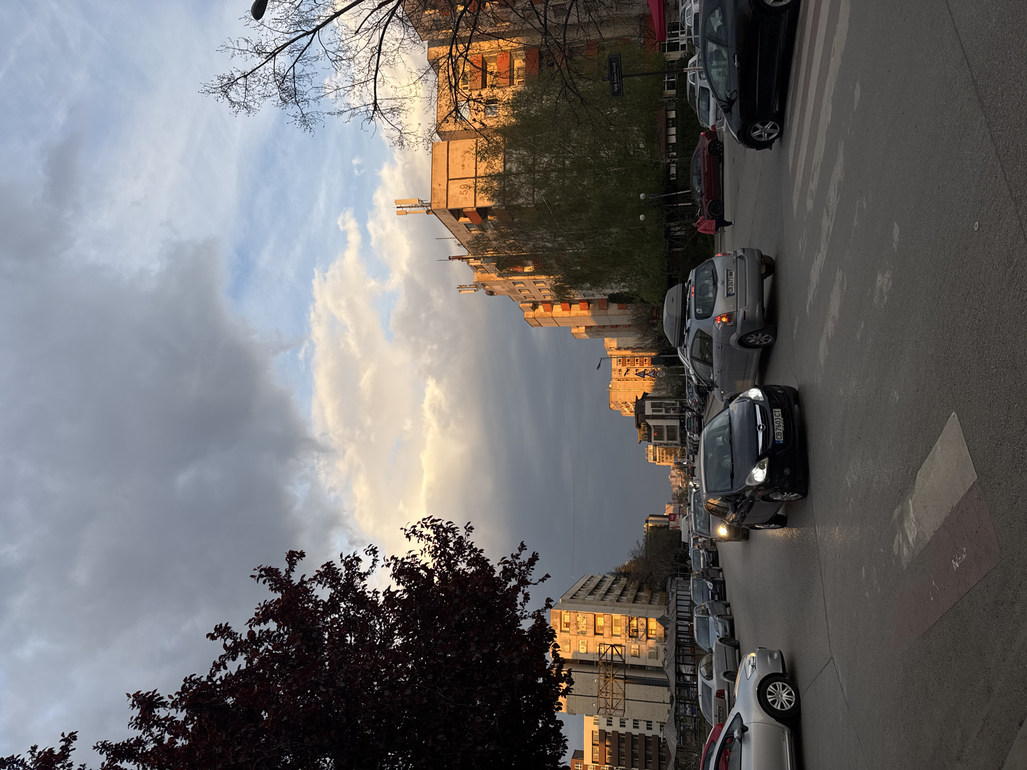 The image presents a vibrant urban street scene in Sofia, Bulgaria, captured during what appears to be the late afternoon or early evening. The sky is dynamic, featuring bright, golden-lit clouds contrasting with darker, overcast grey clouds, creating a dramatic interplay of light and shadow across the city.
The main subjects are the street, numerous cars, and multi-story buildings that line the thoroughfare. Many cars are visible, both parked along the curb and in traffic on the asphalt road. Notable license plates, such as "CB 4263 MM" and "CB 7603 CT," clearly identify the location as Sofia, Bulgaria. Some vehicles have their headlights illuminated, underscoring the approaching dusk. A pedestrian crossing is marked on the road, and a person in a red jacket is partially visible near it, suggesting daily activity.
Buildings on one side of the street are bathed in warm, golden sunlight, highlighting their architectural details, while others appear to be in shadow or under the darker cloud cover. Trees, some with bare branches and one with dense, dark foliage, are prominent in the foreground and along the street, adding natural elements to the concrete landscape.
A key location cue is a street sign, clearly visible on a building, which reads "Проф. Асен Златаров" (Prof. Asen Zlatarov). The overall mood is one of a bustling city transitioning from day to evening under a strikingly varied sky.