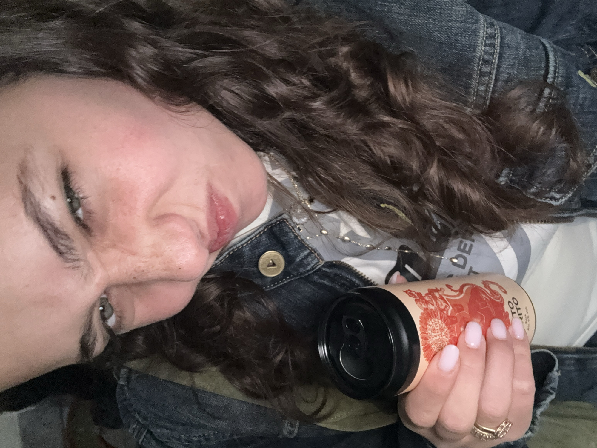 A woman with curly brown hair and fair skin is holding a can with a black top in her right hand. She is wearing a denim jacket over a patterned top. The can appears to be a drink with a design in red. She is also wearing a ring on her finger. The image is taken from a low angle, looking up at the woman. There is no information in the image to suggest it was taken in Milan, Italy.