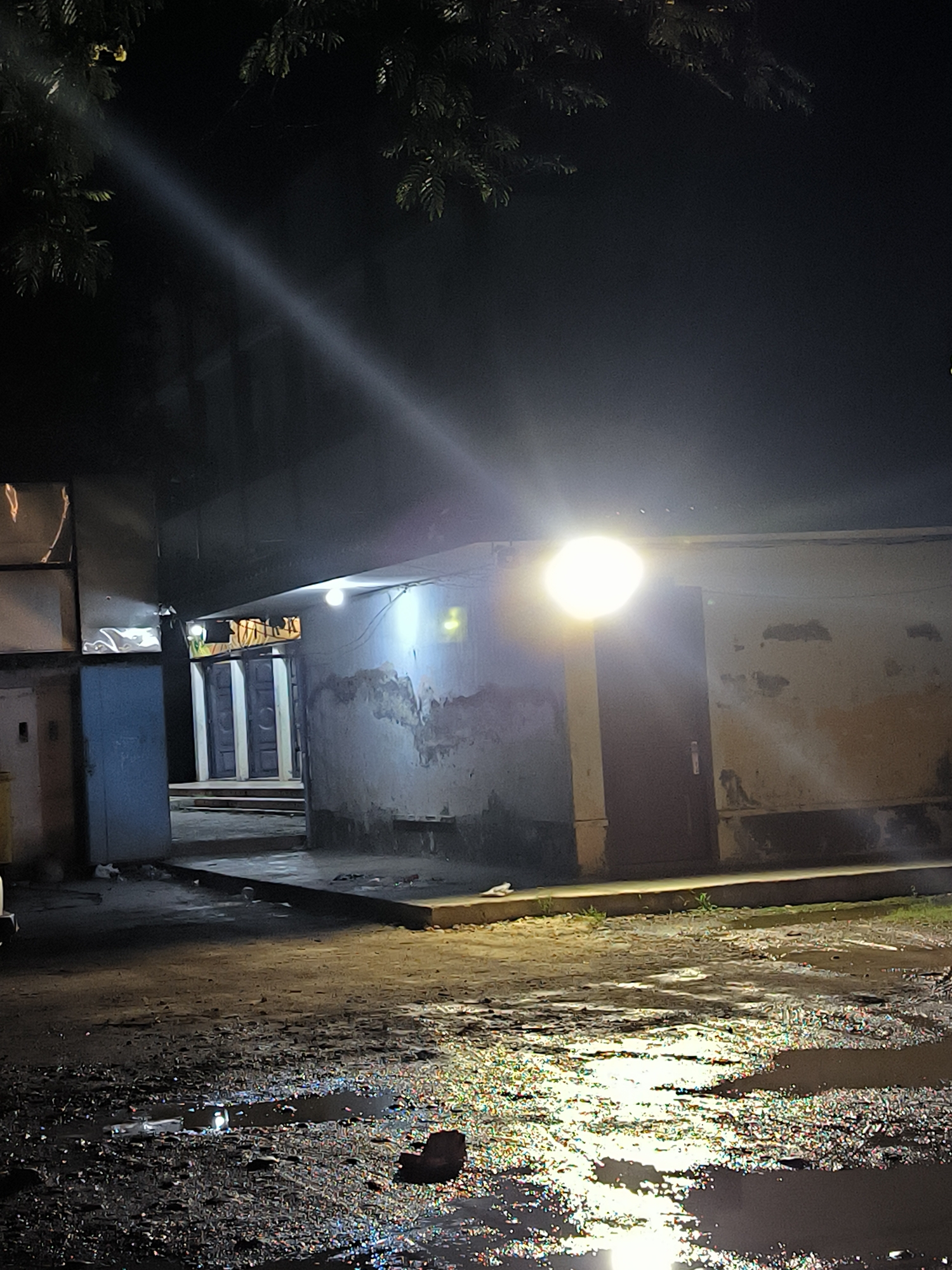 The media file captures a nighttime outdoor scene in Banda Aceh, Indonesia, depicting a path or courtyard between low-rise buildings. The ground in the foreground is wet, muddy, and covered with numerous puddles, brightly reflecting the artificial lights and creating a shimmering, iridescent effect.
On the right side of the image, a simple building features a dark door and is illuminated by a very bright, circular light fixture mounted on its wall, casting a strong glare. To the left, another structure presents a partially open blue door, with a smaller light source visible nearby. Further into the background, a receding pathway leads to an area with multiple dark-colored, paneled doors set within white frames, softly lit from above or within.
Dark foliage from trees or bushes is visible in silhouette against the dark sky in the upper corners of the frame. A prominent, diagonal beam of light cuts across the upper left portion of the night sky, possibly originating from an overhead streetlamp or security light.
No people are visible, and there is no specific activity taking place, suggesting a quiet moment in an urban or semi-urban environment. The overall impression is one of ordinary streets after rain, with artificial lights piercing the darkness. There is no visible text in the image.