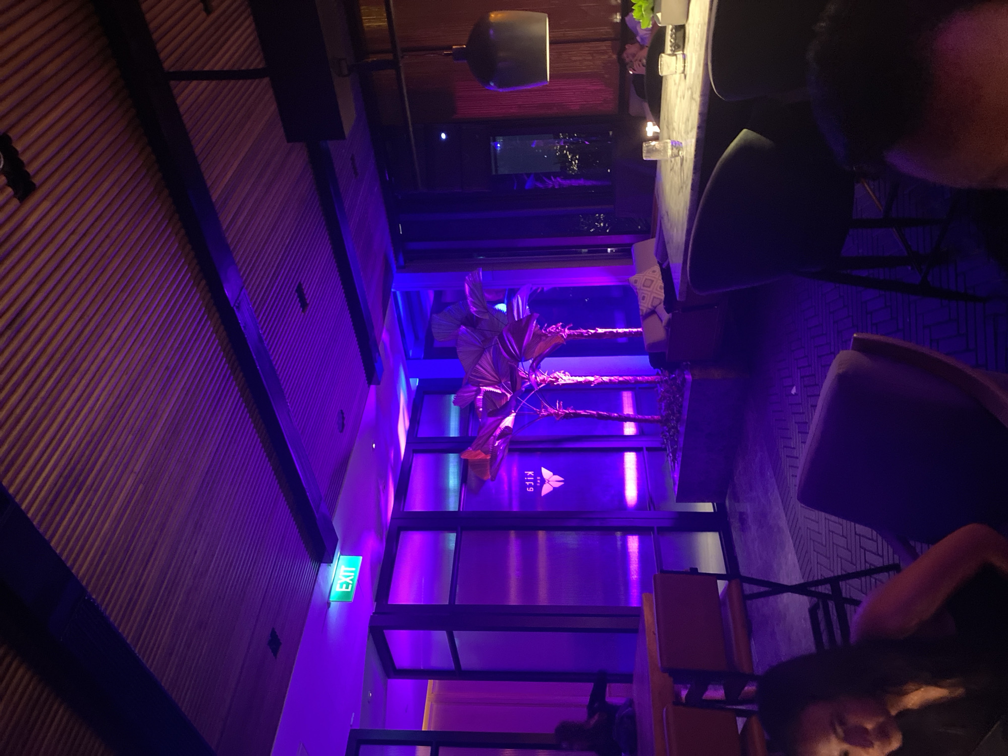 An interior shot of a dimly lit restaurant in Central Jakarta, Indonesia. The space is bathed in purple and pink neon lighting, illuminating a modern dining area. A green "EXIT" sign is visible above a doorway on the left. The room features sleek wooden ceilings, plush seating, and decorative palm trees. A marble-topped bar with stools is present, with glasses and other barware on its surface. Patrons are seated at tables, some of whom are partially visible. The overall ambiance suggests a trendy and upscale establishment.