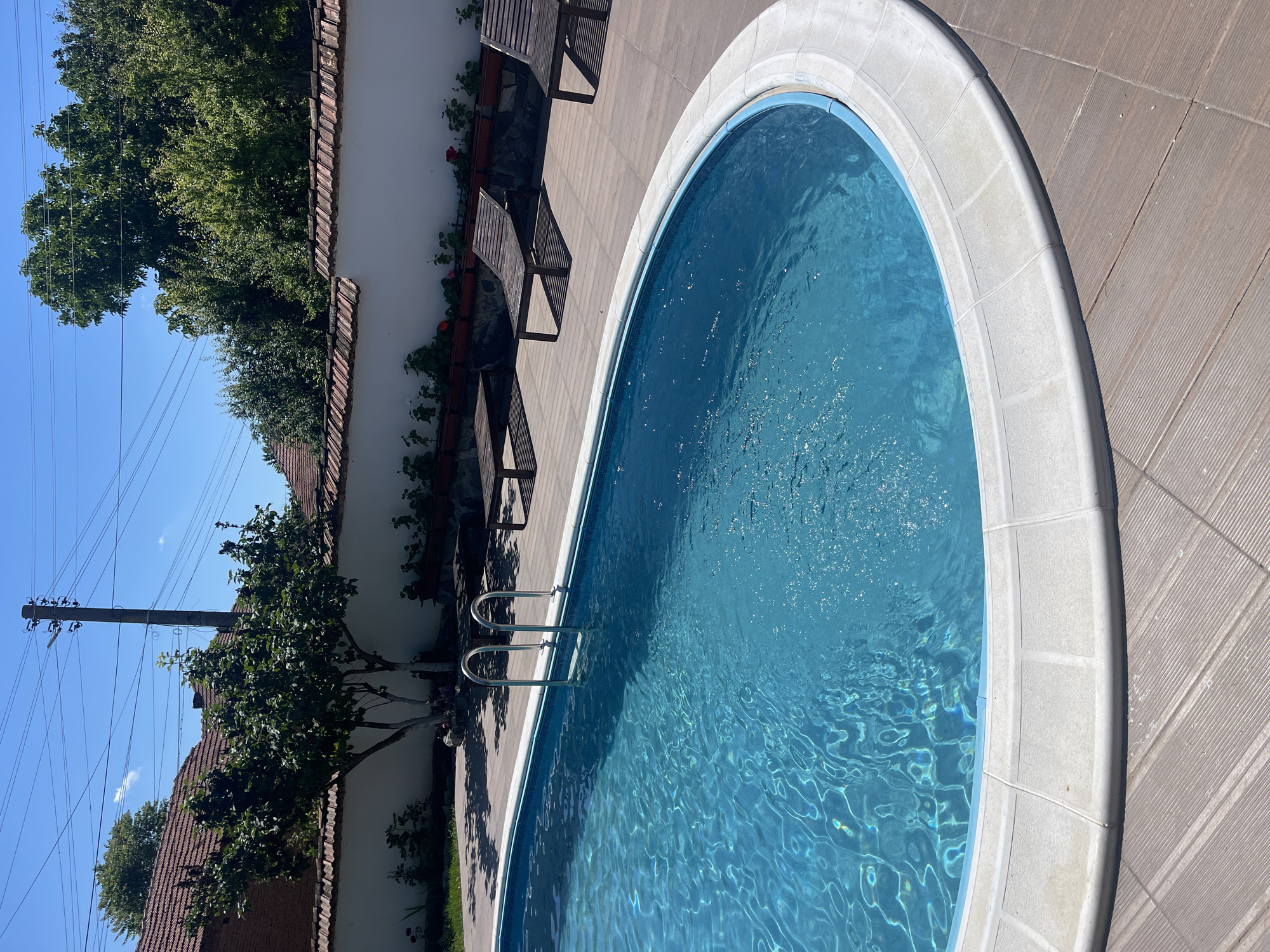 A bright, sunny day illuminates an outdoor swimming pool with a blue tiled interior and a white coping edge. A silver ladder extends into the pool from the wooden deck surrounding it. Several wooden lounge chairs are placed on the deck, casting shadows on the ground. Behind the pool area, a white wall topped with red-tiled roof separates the yard from a residential area. A leafy tree with white-painted trunks stands near the wall, and more trees and a tiled roof are visible further back. Power lines and a utility pole are visible against the clear blue sky. The image appears to be taken from a slightly elevated position, looking down and across the pool area. The location is the town of Totleben, Bulgaria.