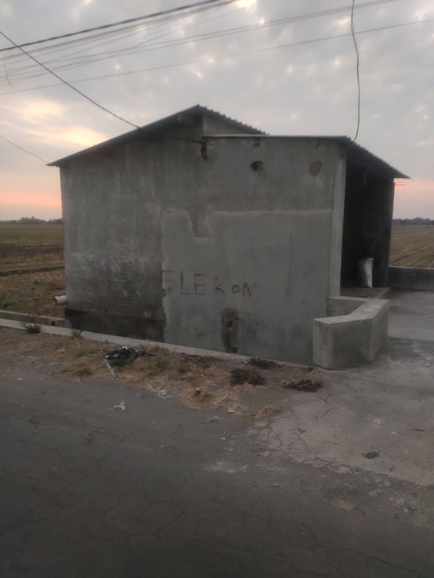 The image depicts an exterior view of a small, unfinished concrete structure, likely a shelter or a small building, situated outdoors in Bongkot, Indonesia. The structure has a corrugated metal roof and appears to be built from concrete blocks or poured concrete, with some visible imperfections and marks on its walls. The word "ELEKON" is spray-painted in red letters on the front wall of the structure. Adjacent to the building, there is a low concrete wall or seating area. The ground surrounding the structure is a mix of cracked concrete pavement, dry grass, and some scattered debris. In the background, an open field with agricultural rows is visible, and a network of overhead power lines stretches across the cloudy sky. The lighting suggests it is either early morning or late afternoon, with a soft, diffused light illuminating the scene. There are no people visible in the image.