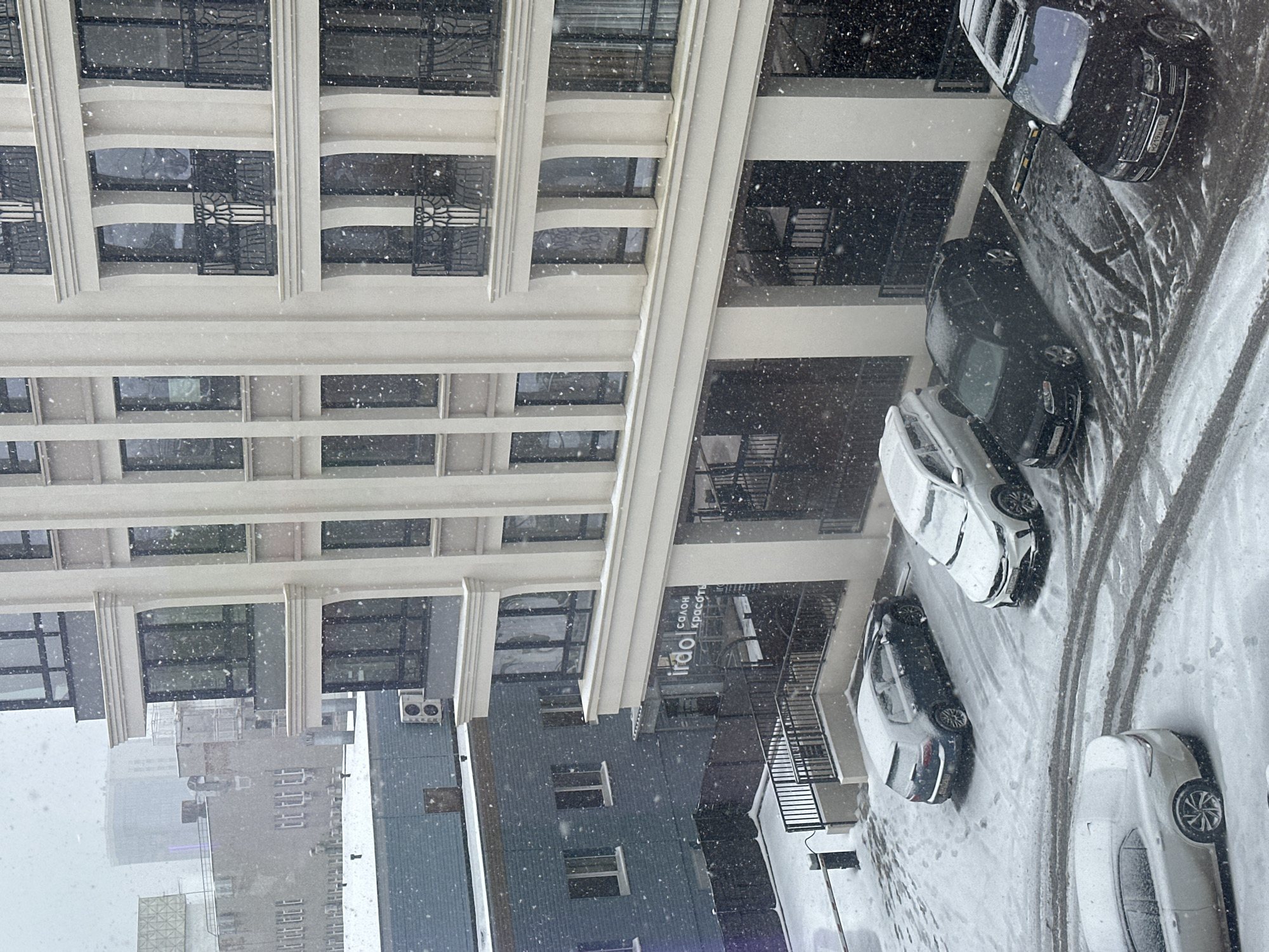 The image depicts a snowy day in what appears to be an urban environment, potentially in Perm, Russia, based on context. A multi-story building with a light-colored facade and numerous windows is prominently featured. Parked cars are visible along the street beside the building, partially covered in snow. Other buildings and structures can be seen in the background, suggesting an urban setting. The environment is characterized by snowfall and associated accumulation on surfaces.