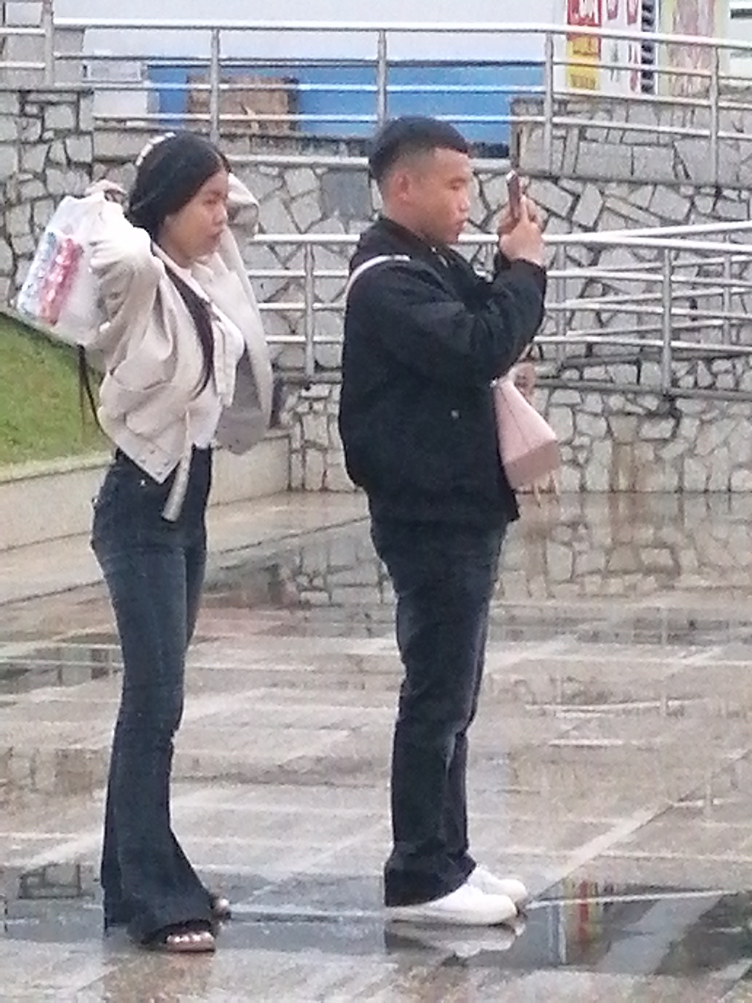The image depicts a young Asian couple standing on a wet, paved surface, likely outdoors in Da Lat, Vietnam. The woman, on the left, wears a white top, a light-colored jacket, and dark, flared jeans. She carries a white and pink bag over her left shoulder. The man, on the right, is dressed in a black jacket and dark pants. He holds a smartphone in his right hand, appearing to take a picture or record a video. A light pink handbag rests near his left hip. In the background, a stone wall and metal railings are visible, suggesting a public area such as a park or plaza. The ground is reflective due to recent rain.