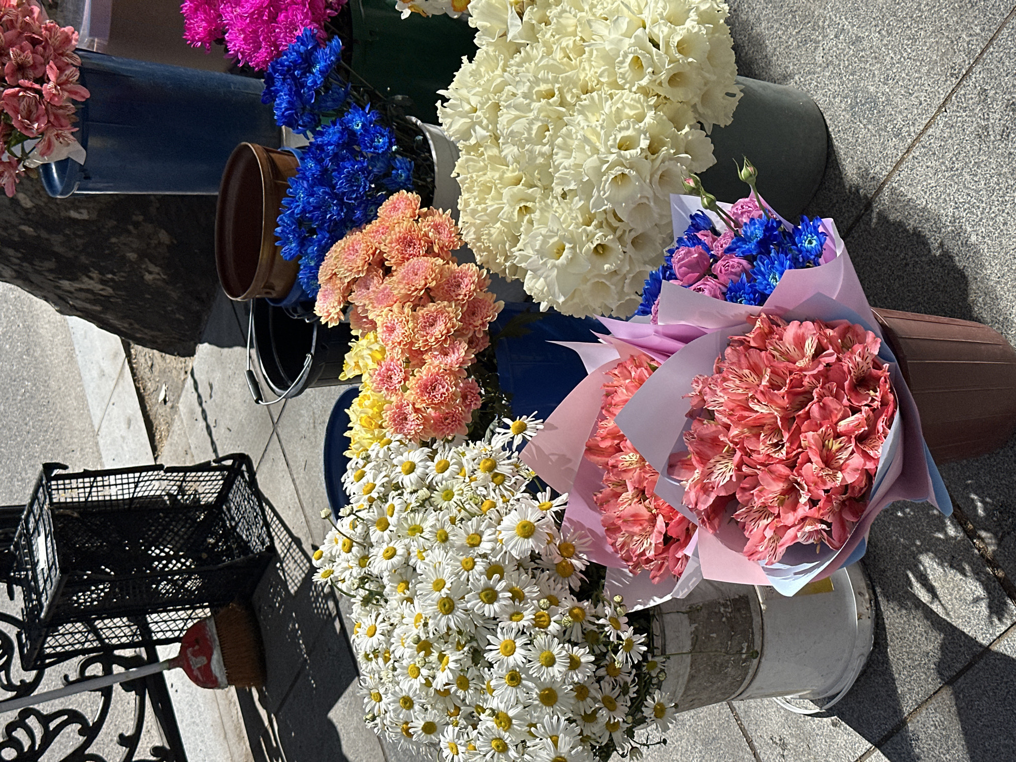 The image depicts a flower stall in Tbilisi, Georgia, situated on a paved area. Assorted flower bouquets and bunches are displayed in various buckets and containers. The flowers include white daffodils, blue and pink chrysanthemums, orange chrysanthemums, daisies, and pink alstroemeria bouquets wrapped in colored paper. A black metal basket and a broom lean against a wrought iron bench next to the flowers. The background includes a building, trees, and shadows indicating a sunny day.