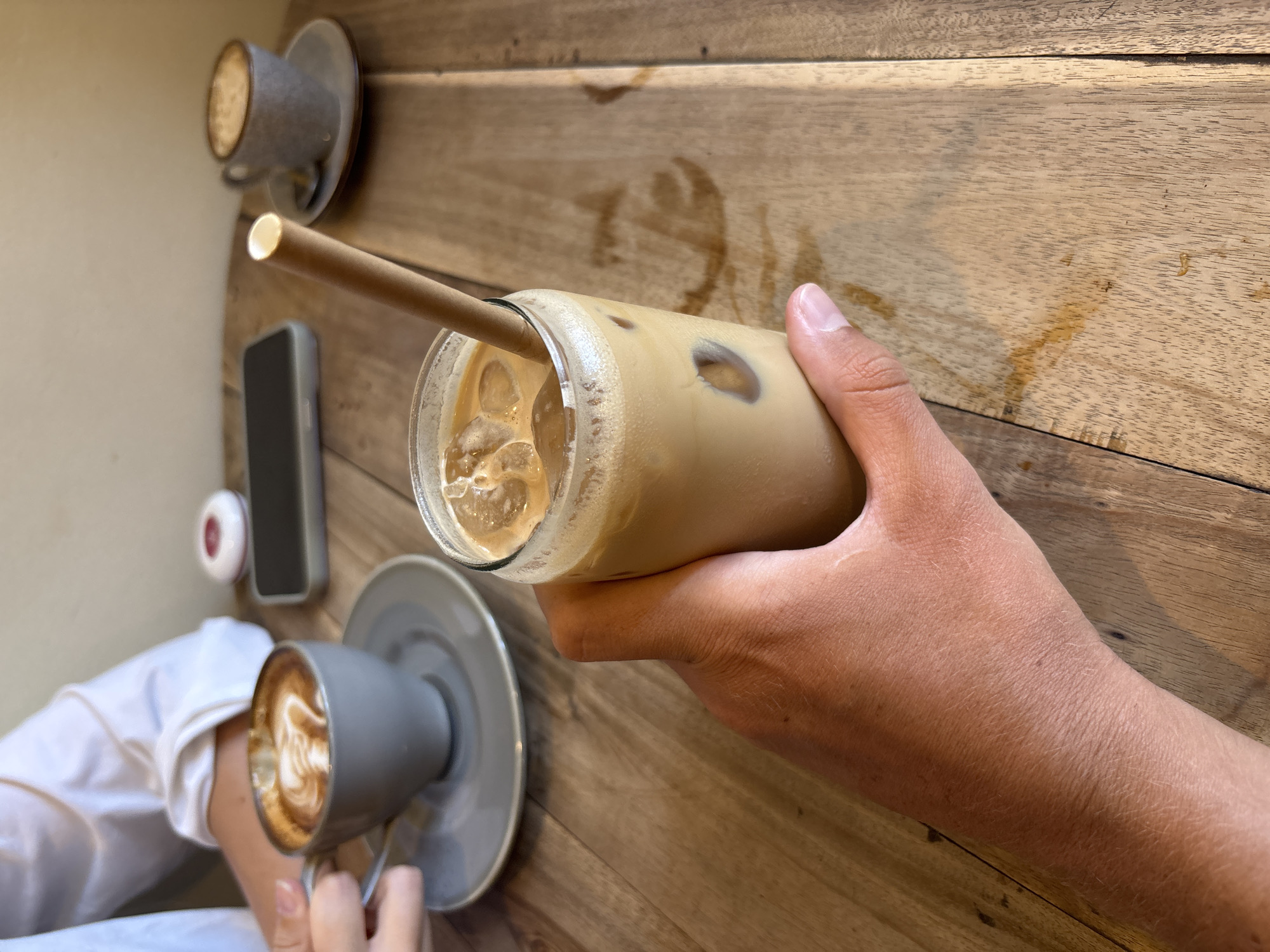 Captured from an overhead perspective on a wooden table, the photograph features several elements that suggest a relaxed, possibly café-like setting. A fair-skinned individual's hand is seen holding a tall, transparent glass filled with ice and a light-colored beverage, likely a coffee drink. A paper straw extends from the glass. Adjacent to this, two grey cups on matching saucers contain coffee beverages, hinting at the presence of companions. A mobile phone and what appears to be a white earbud case also sit on the table.
Given the specification of Kuta Selatan, Indonesia, it is conceivable that this scene is situated in a café or similar establishment in that area. However, no definitive elements within the image confirm this location.