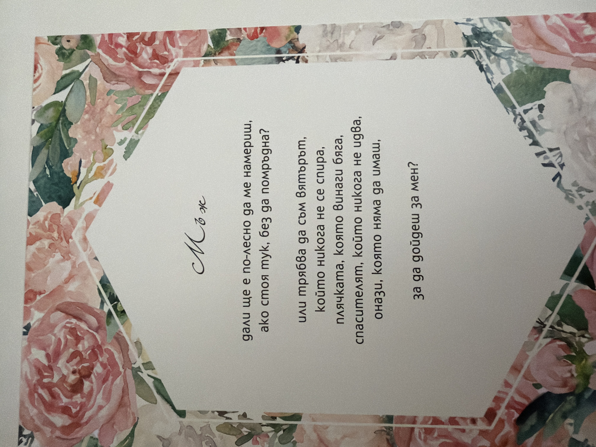 The image shows a part of a greeting card with floral watercolor designs on the borders and a poem written in the center. The poem is in Bulgarian. The central design is a white hexagonal shape with text inside. The text reads: "Мъж / дали ще е по-лесно да ме намериш, / ако стоя тук, без да помръдна? / или трябва да съм вятърът, / който никога не се спира, / плччката, която винаги бяга, / спасителят, който никога не идва, / онази, която няма да имаш, / за да дойдеш за мен?" The floral watercolors depict pink and white roses with green leaves. The image quality is slightly grainy. There is no information about the location within the city of Pazardzhik, Bulgaria.