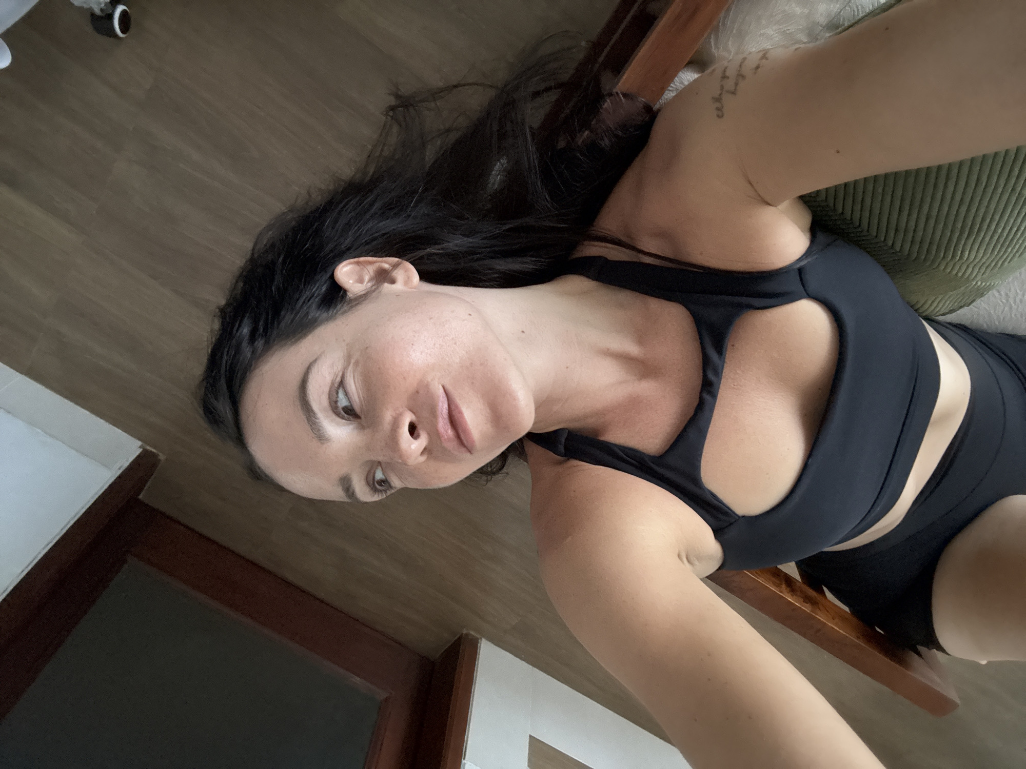 A woman with dark hair and fair skin is captured from a high angle, looking to her right. She is wearing a black sports bra with a cutout design and black shorts. Her right arm rests on a textured green cushion, and a tattoo is visible on her upper right arm. The background consists of wooden flooring and a portion of a window frame with dark wood trim. The scene appears to be indoors, likely in a residential setting in North Kuta, Indonesia.