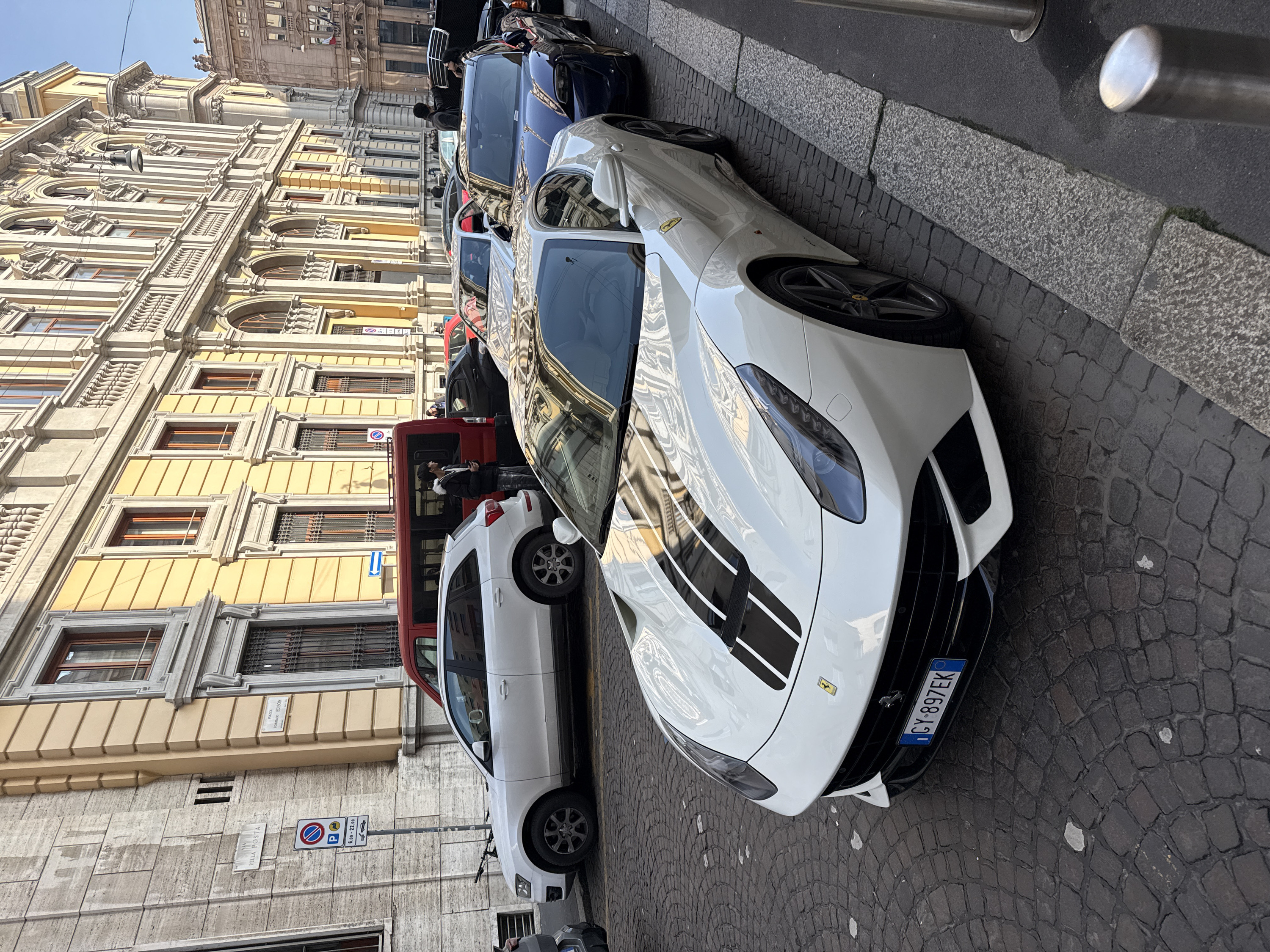 The image presents an urban street scene in Milan, Italy, at the intersection of Via della Posta and Piazza Tommaso Edison, under natural daylight conditions.
In the foreground, a white Ferrari, identified by its two central black racing stripes and Italian license plate "CY-897EK," is parked on dark grey cobblestones. Its front fascia features prominent headlights and black multi-spoke wheels.
Behind the Ferrari, a sequence of vehicles is parked parallel to the curb. Immediately following is a light grey Porsche. Subsequently, a dark blue car, likely a Porsche Taycan, is parked with its rear trunk open. A male figure in a dark jacket is observed leaning into the open trunk, interacting with a female figure possessing dark hair. Further left in the background, a white Audi Q5 SUV is parked on the cobblestones, carrying a bicycle on its roof rack. Adjacent to the Audi, a red van is parked, also equipped with a bicycle on its roof.
A young male individual, dressed in a black puffer jacket with a fur-lined hood and light jeans, stands near the red van, holding and observing a white smartphone.
The street is flanked by multi-story buildings. The prominent building on the right is painted yellow and features ornate light-colored architectural details, including decorative window surrounds, balustrades, and pilasters. The building on the left exhibits a light grey, stone-block facade. Street signage clearly identifies "VIA DELLA POSTA" and "PIAZZA TOMMASO EDISON." A regulatory sign indicates no parking between "6.00 - 22.00." The street surface is composed of dark grey cobblestones, separated from an asphalt sidewalk by a light grey granite curb.