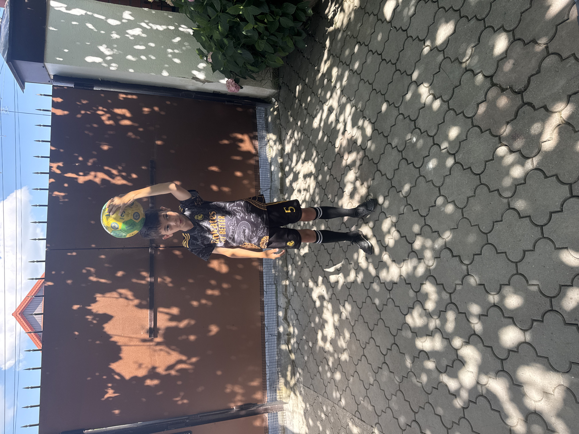 A young boy stands outdoors on a paved patio in front of a dark brown gate. He is wearing a black and yellow soccer jersey with dragon graphics and white text, black shorts with the number "5" in yellow, black socks pulled up to his knees, and black soccer cleats. The boy is balancing a yellow and green soccer ball on top of his head with his right hand resting on the ball. The sun casts dappled shadows from tree leaves onto the gate and the patio. To the right of the gate is a white pillar with a darker trim, and a bush with pink flowers is visible next to it. The sky is blue with some white clouds.