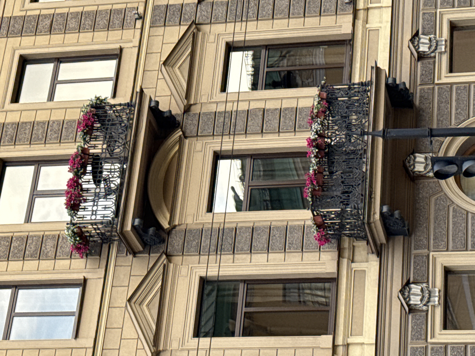 The image shows a close-up view of a building facade in Saint Petersburg, Russia. The building appears to be old, with ornate architectural details. The facade is a light beige color and features several windows with dark frames. Two balconies with decorative wrought-iron railings are visible, each adorned with hanging planters filled with vibrant pink and white flowers. The balconies are positioned on different levels of the building.
Above the windows, there is a band of decorative square panels with an intricate pattern. The building also has vertical grooved elements and triangular decorative features. In the foreground, a traffic light structure with dark gray metal components is visible, extending horizontally across the frame. The lighting suggests it might be daytime, possibly overcast, as there are no strong shadows. The overall impression is one of urban architecture with touches of floral beauty. No people are visible in the image, and no text is present.