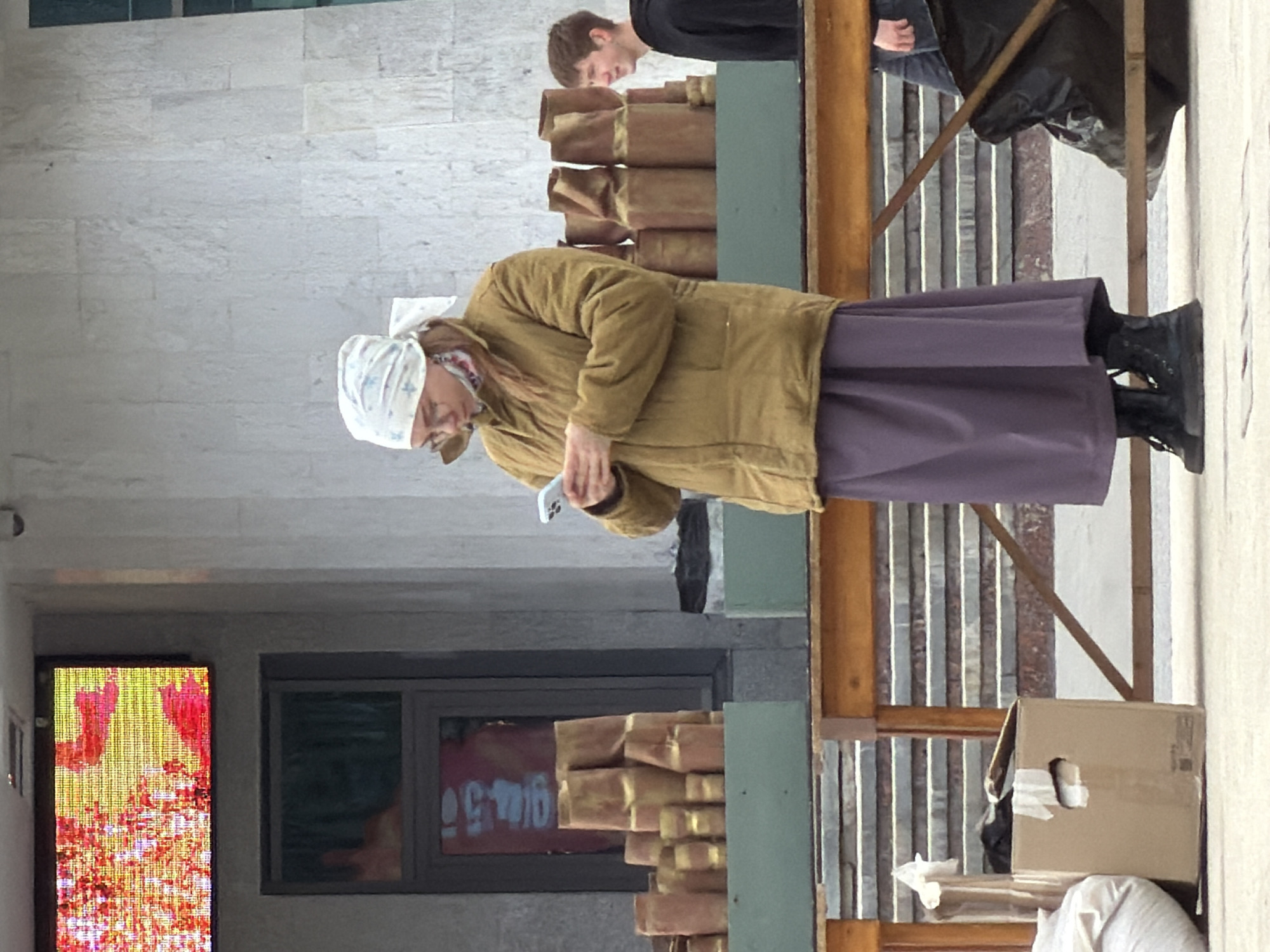An eye-level, outdoor shot depicts an older woman in a light-brown jacket, a long, purple skirt, and black boots, standing near a table of stacks of brown, wrapped items outside a building with a lit window. The woman is holding a phone. Behind the woman, a young man with short brown hair is standing near the same table. A cardboard box and other objects are at the edge of the frame. The building, assumed to be in Izhevsk, Russia, has a lit window with colorful designs.