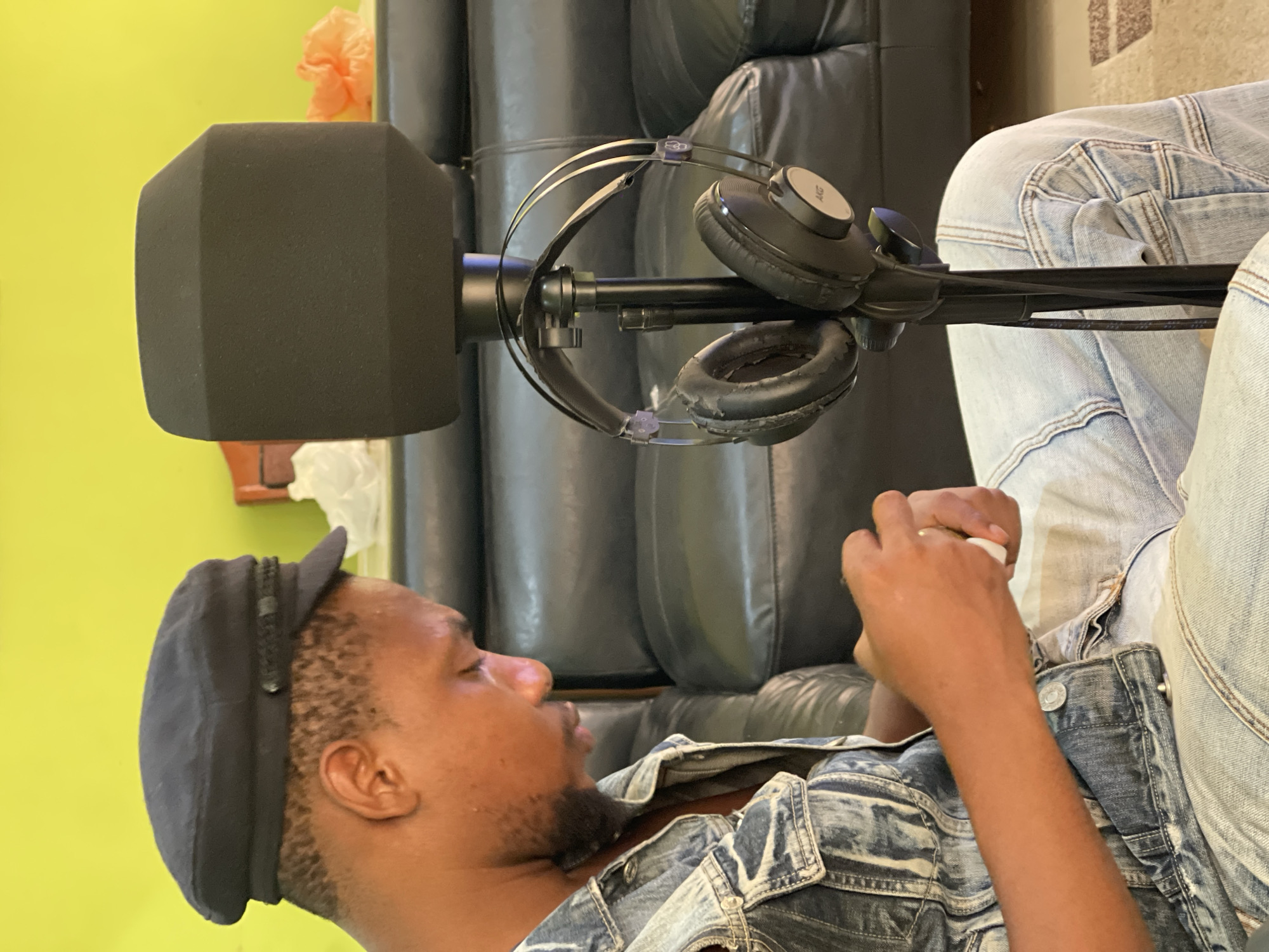 The image shows a dark-skinned male, likely in his late 20s or early 30s, wearing a dark cap and a denim jacket. He appears to be reclining, possibly on a black leather couch. A microphone with a large black windscreen is positioned close to him. A pair of over-ear headphones hangs from the microphone stand. He seems to be holding something small and white in his hands, possibly a piece of paper or a small object.
The background consists of a yellow wall, and a partial view of what looks like the wall to floor finish.
Based on the metadata, the image was taken in Abuja, Nigeria. The scene suggests a casual, indoor setting. It could be a home recording setup or a relaxed atmosphere. There is no specific information to define precise location in the city.