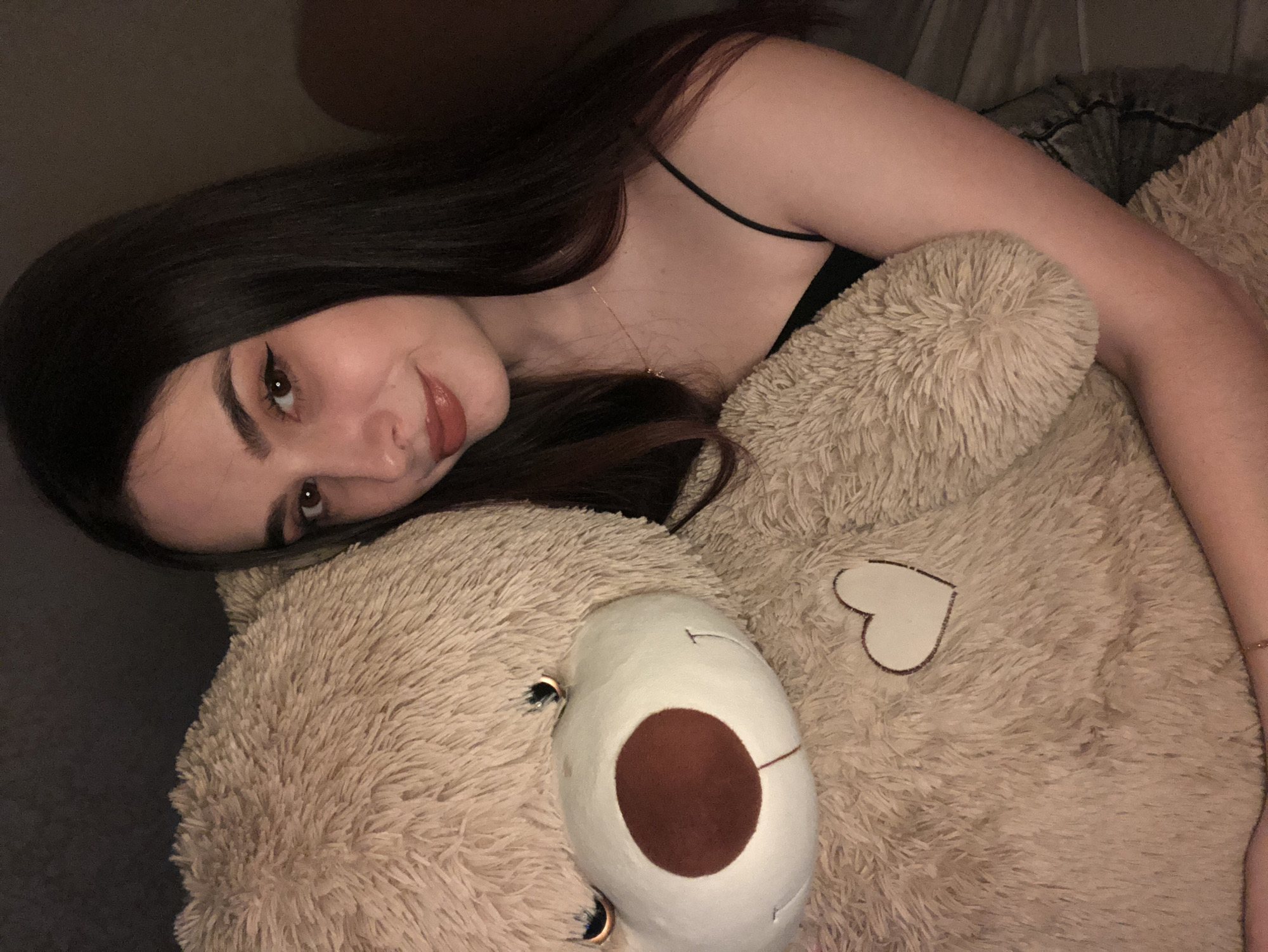 A young woman with long, dark hair and wearing a black tank top is lying on top of a large, light brown teddy bear. The teddy bear has a large white snout with a brown nose and small, dark eyes. A white heart-shaped patch is sewn onto the bear's fur. The woman is looking at the camera with a slight smile. The background is dark, suggesting the photo was taken indoors. The location is not discernible from the image, and there is no evidence to suggest it is Noviy, Russia.