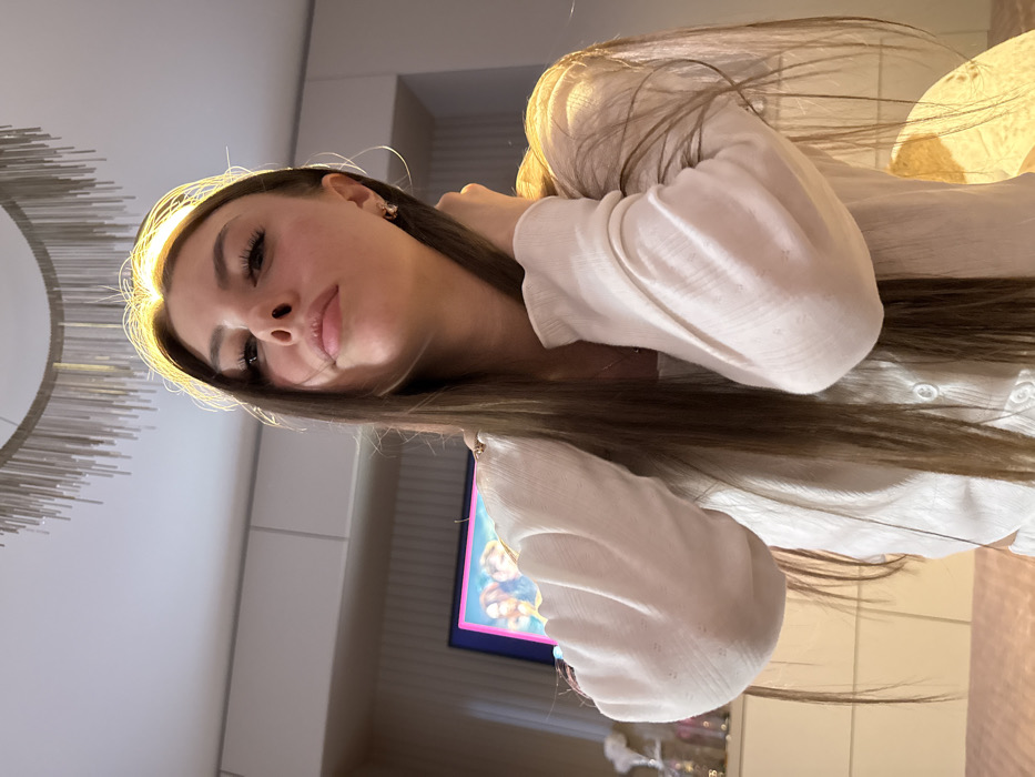The scene is an indoor environment in the city of Dubovaya, Russia. A young adult female is centrally positioned, captured from a low-angle perspective looking slightly upwards towards the camera. She possesses long, straight, dark brown hair, which flows over her shoulders and chest, with a distinct bright golden halo illuminating the top of her head. She is wearing a light-colored, ribbed, long-sleeved top with two visible white buttons on the torso. Her right hand is raised, with fingers partially obscured within her hair near the right side of her neck.
The background features a light-colored ceiling from which a modern chandelier, composed of numerous suspended metallic rods, hangs. Below the ceiling, light-toned wall panels or cabinetry are visible. To the lower left, a television screen displays a colorful image dominated by pink, blue, and orange tones. Below the television, on a flat surface, several clear glass containers hold white, feathery decorative elements. To the lower right, an off-camera light source, likely a lamp with a textured, light-colored shade, emits a warm glow. The immediate foreground reveals a soft, pink, textured surface, possibly upholstery.