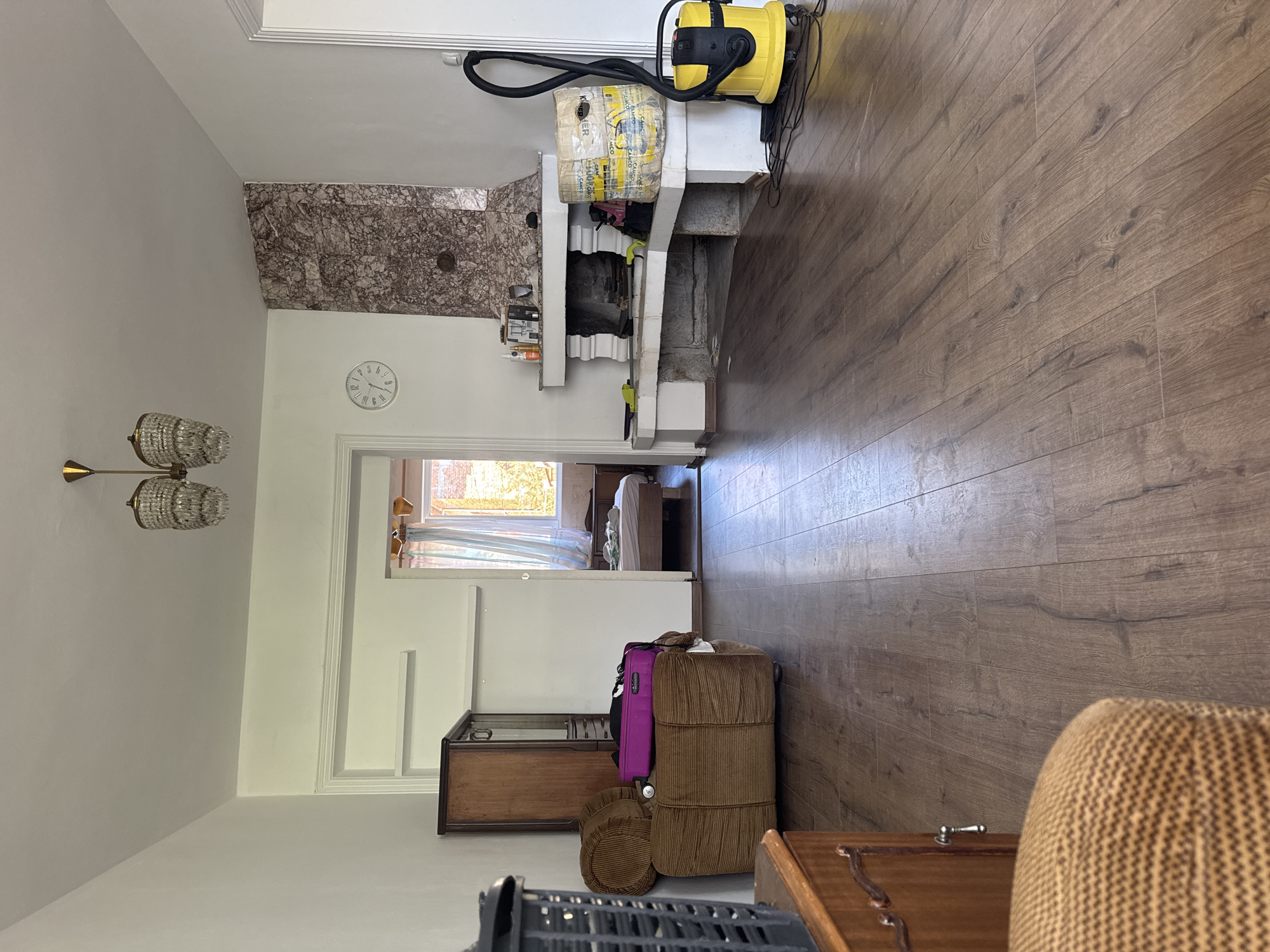 The image depicts an interior scene of a living room. On the left, there is a dark wooden cabinet and a brown armchair with purple luggage on it. A black basket is partially visible in the foreground. In the center and to the right, a large fireplace with a marble surround is prominent. Next to the fireplace, a yellow vacuum cleaner and a large roll of material wrapped in plastic are present. The room has wooden flooring and white walls. Through an open doorway in the background, a bedroom with a bed and window is visible. The overall impression is of a lived-in space that might be undergoing some form of reorganization or cleaning. The specific location in the city of Plovdiv, Bulgaria, cannot be determined from the visual information provided in the image.