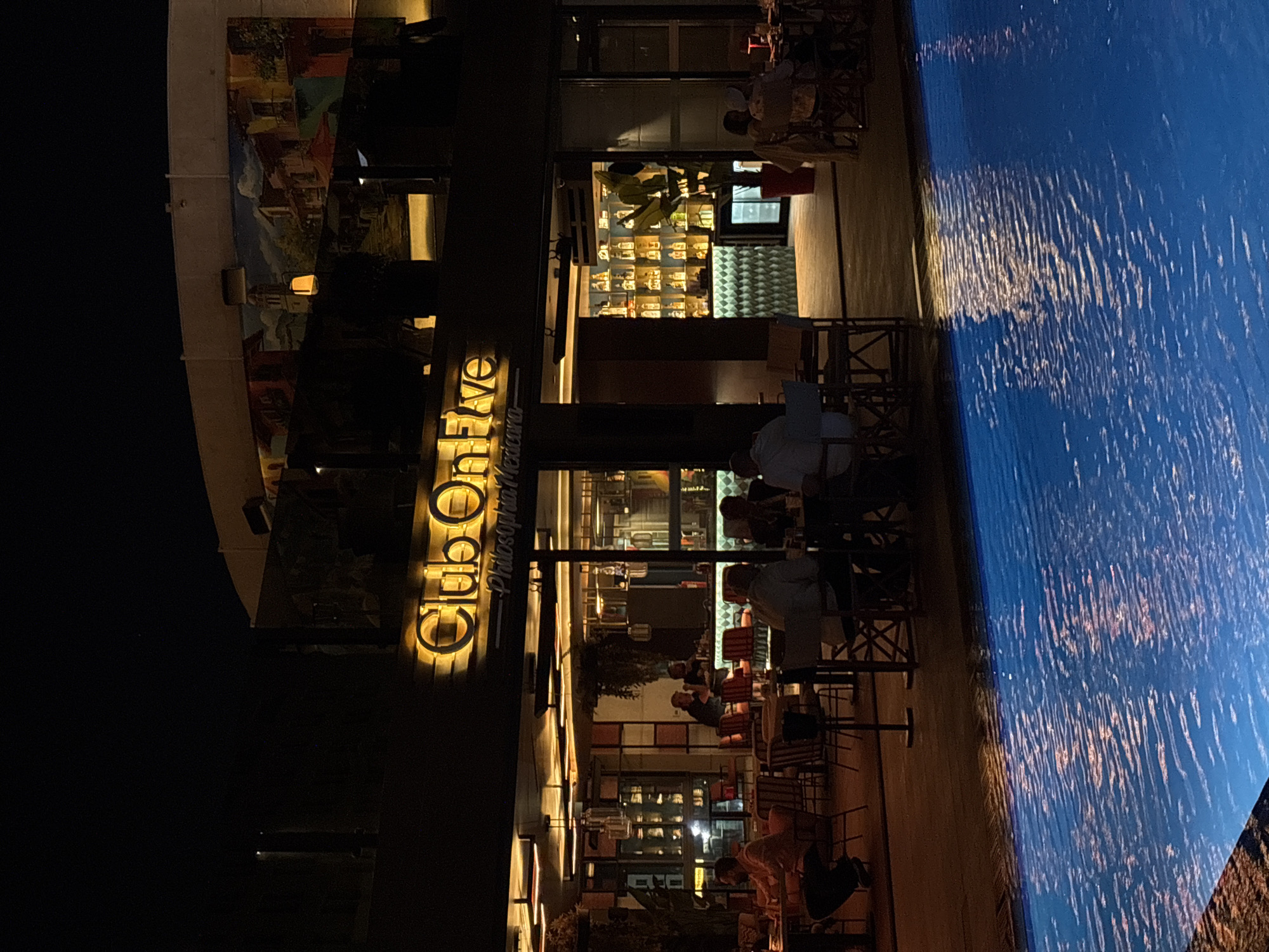 The image depicts the exterior of "Club On Five" restaurant and bar at night, located in Amman, Jordan. A brightly lit sign with the text "Club On Five" and "Philosophia Mexicana" is prominently displayed. The restaurant features an outdoor seating area with tables and chairs, some of which are occupied by patrons. Behind the outdoor seating is the restaurant's interior, with a bar area stocked with bottles and illuminated shelving. A swimming pool is visible in the foreground, with reflections of the restaurant's lights shimmering on its surface. The overall atmosphere is that of a lively evening establishment.