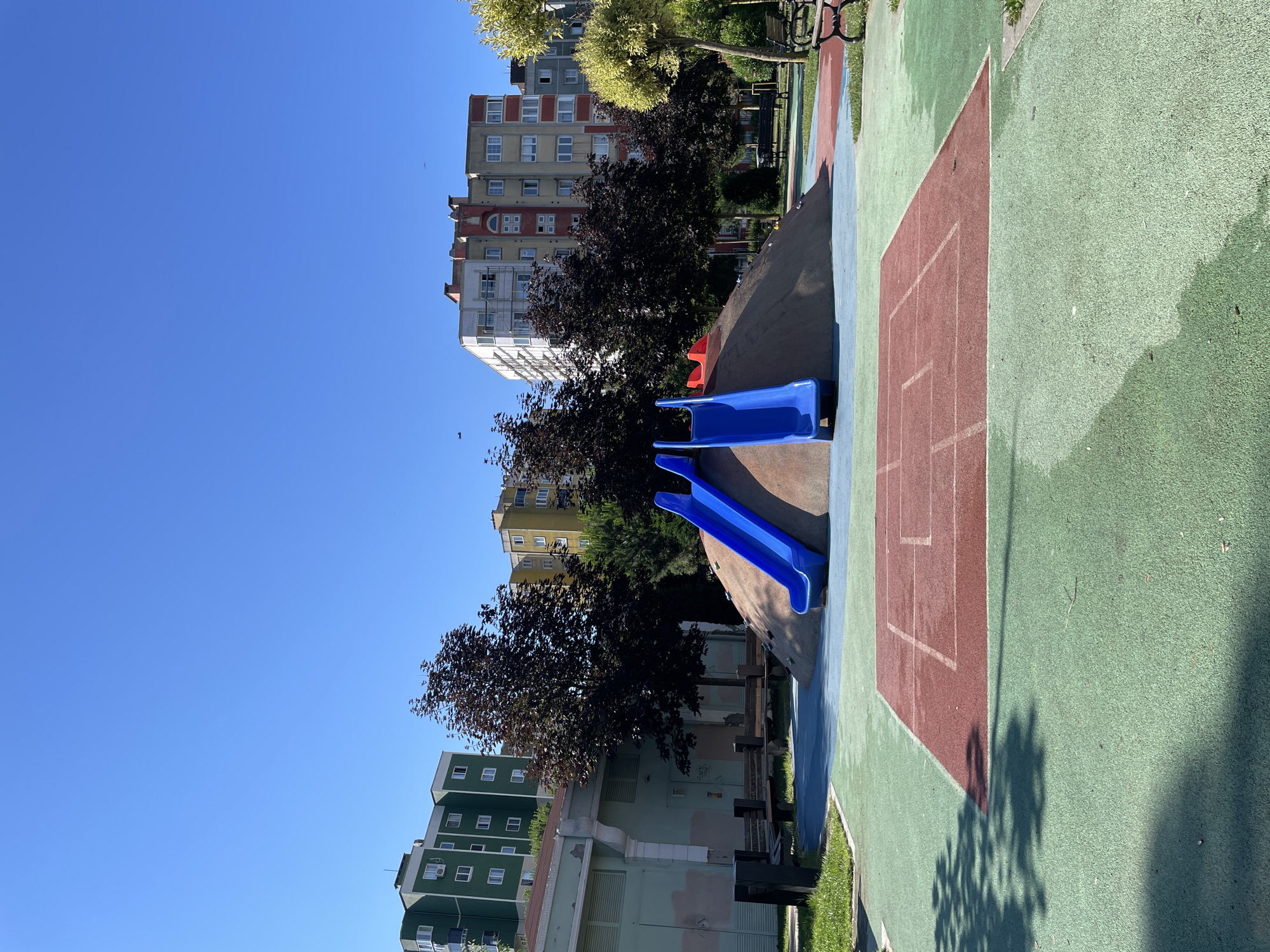 The image shows a children's playground in Başakşehir, Turkey, on a clear day with a bright blue sky. In the foreground, there is a green rubberized surface with a red rectangular area marked with white lines, resembling a game court. Behind this, a sand-covered mound features two blue slides. Trees with dark foliage are situated behind the mound, and in the background, several apartment buildings of varying colors, including green, yellow, and beige, are visible. Benches are placed around the playground area. The scene is devoid of people.
