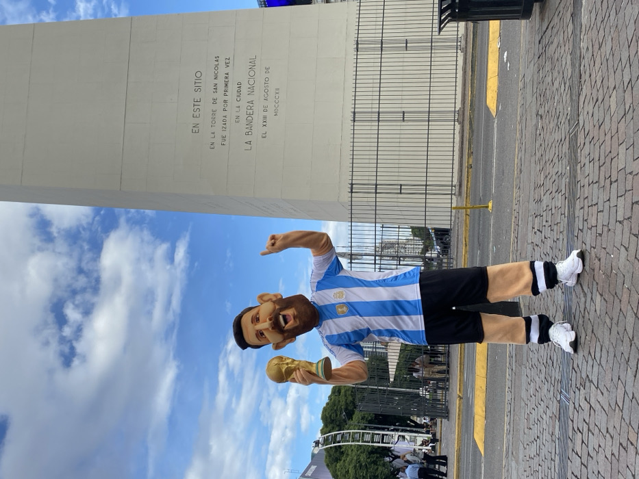 An individual in a Lionel Messi mascot costume stands centrally on cobblestone pavement, facing the viewer, in front of the Obelisco de Buenos Aires. The costume features an oversized head with a smiling, bearded likeness of Messi, an Argentina national football team jersey with blue and white stripes and a FIFA World Cup winner's badge, black shorts, tan-colored leg and arm coverings, and white athletic shoes. The mascot holds a gold replica of the FIFA World Cup trophy in its left hand and points its right index finger upwards.
Behind the mascot, to the right, is a prominent section of the white Obelisco de Buenos Aires, located in the Plaza de la República, Buenos Aires, Argentina. An inscription is visible on its surface, reading "EN ESTE SITIO EN LA TORRE DE SAN NICOLAS FUE IZADA POR PRIMERA VEZ EN LA CIUDAD LA BANDERA NACIONAL EL XXIII DE AGOSTO DE MDCCCXII." A black metal fence runs horizontally across the mid-ground, separating the immediate foreground from the obelisk. To the left background, a large Ferris wheel-like structure, trees, and several indistinct figures are visible. A street with a yellow painted curb lies between the foreground and the background elements. A dark trash receptacle is positioned in the far right foreground. The scene is outdoors under a partly cloudy blue sky with bright daylight.