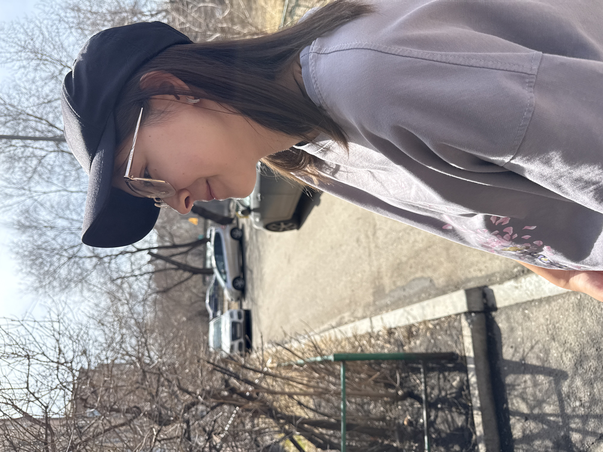 A woman in a black baseball cap and glasses looks down. She is wearing a light gray shirt with a floral design. The background includes a street with cars, bare trees, and a building. The location is most likely a balcony or elevated position overlooking a street in Krasnoyarsk, Russia, due to the urban setting and typical architecture.