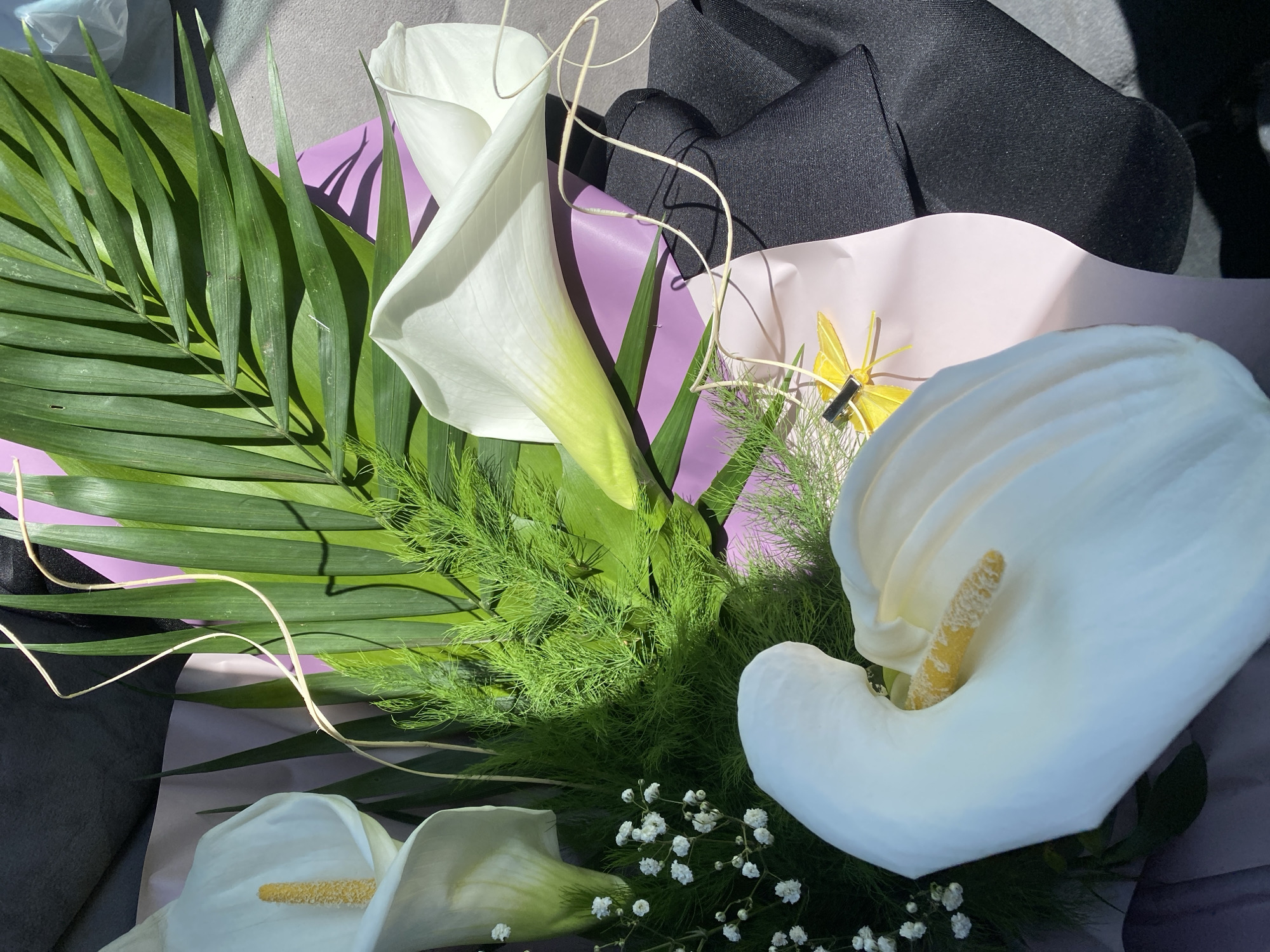 The image shows a bouquet of white calla lilies with green foliage, arranged with decorative paper and string. A yellow butterfly accessory is visible. A black garment or piece of fabric is partially visible behind the bouquet. The location is not identifiable from the image.