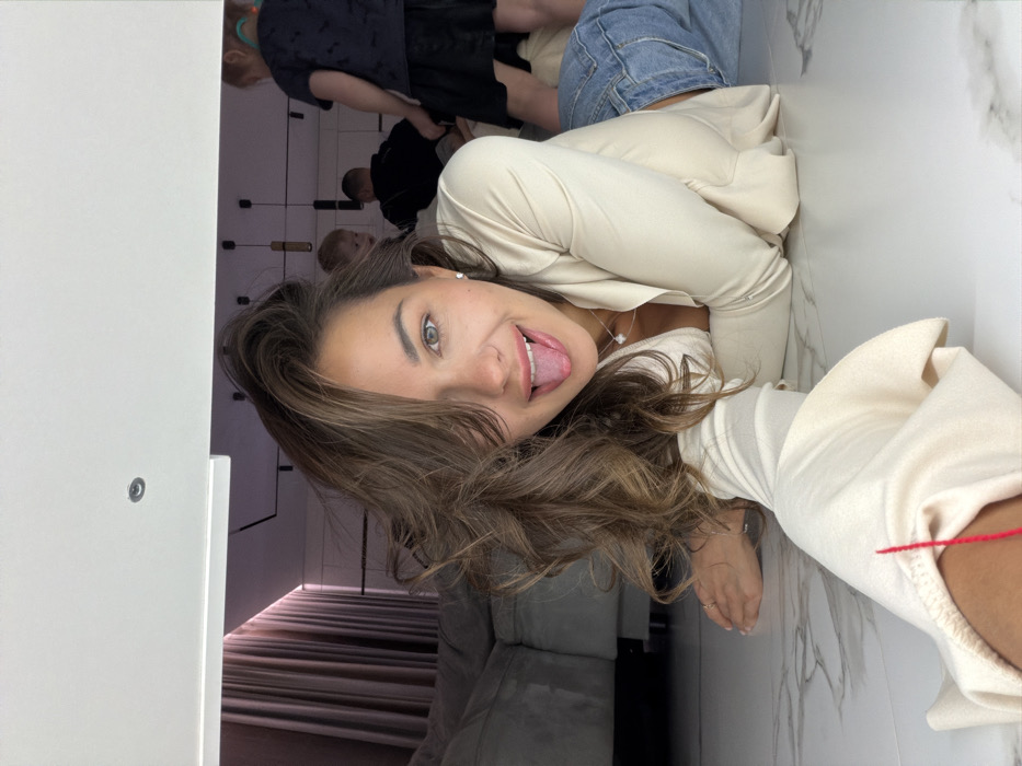 A brunette woman sticks out her tongue, smiling at the camera. She wears a tan long-sleeved shirt and necklace, posed against a white marble-look wall and floor. In the background, a blurry group of people stand, and a striped carpet is visible near a white sofa. A white door is to the left.
