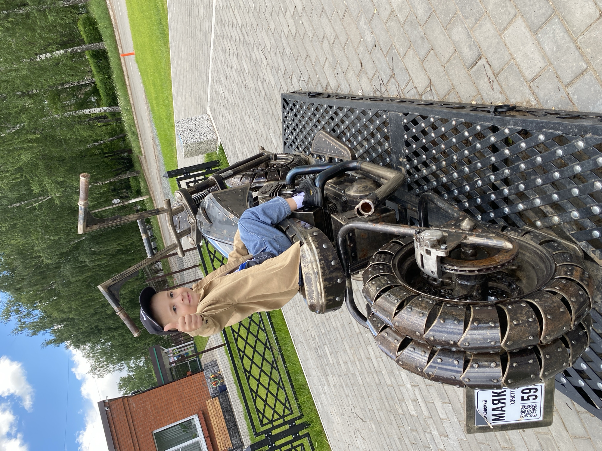 A young boy, wearing a tan jacket, blue jeans, black shoes, and a black baseball cap, is seated on a motorcycle sculpture made of metal. He is giving a thumbs-up. The motorcycle is situated on a paved area. In the background, there are trees, grass, a fence, and a building with a brick facade. The motorcycle's license plate reads "МАЯК" and "59," which indicates the city of Chaikovsky in the Perm Krai region of Russia.