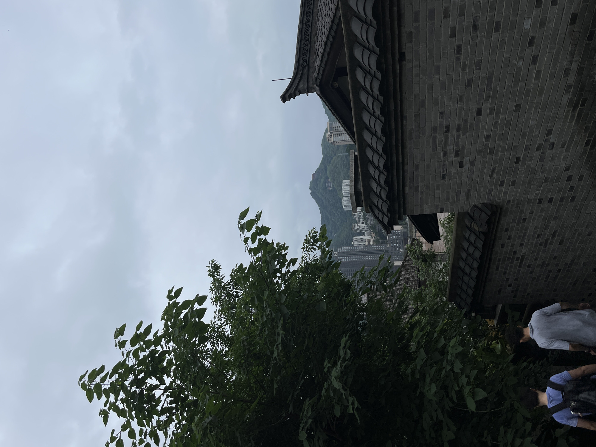 The image presents an exterior shot in Yuzhong District, China, under an overcast sky. The focal point includes a building with traditional East Asian architecture, characterized by dark gray brickwork and a tiered roof with downturned eaves. The building appears to be elevated, providing a view of a mountainous area in the background dotted with high-rise buildings. Green foliage from nearby trees partially obscures the view of the cityscape. In the lower right corner, there are two people, one wearing a gray shirt and another a blue shirt with a backpack. They are situated in proximity to the building, suggesting they may be tourists or visitors.