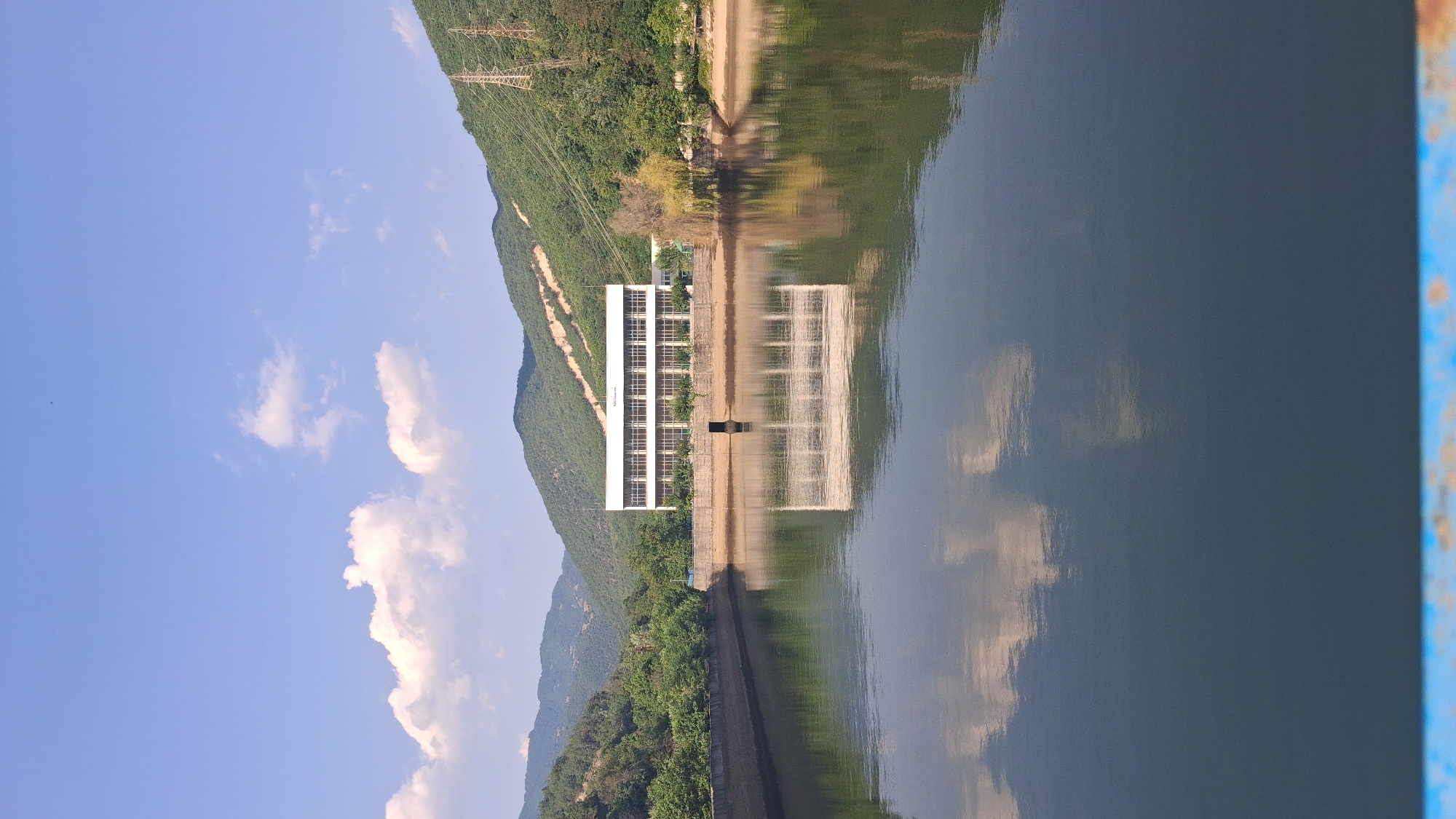 The image depicts a serene landscape dominated by a large, calm body of water reflecting the surrounding environment under a clear blue sky with scattered white clouds. A long, rectangular building with numerous windows stands prominently on the far bank of the water, its facade also reflected in the water. The building appears to be a dam control or administrative structure, likely part of the Gorno Spanchevo Dam in Bulgaria, given the context. Green, tree-covered hills rise behind the building, forming a natural backdrop. The water's edge is lined with a concrete embankment. The overall scene is one of natural beauty and functional infrastructure coexisting harmoniously.