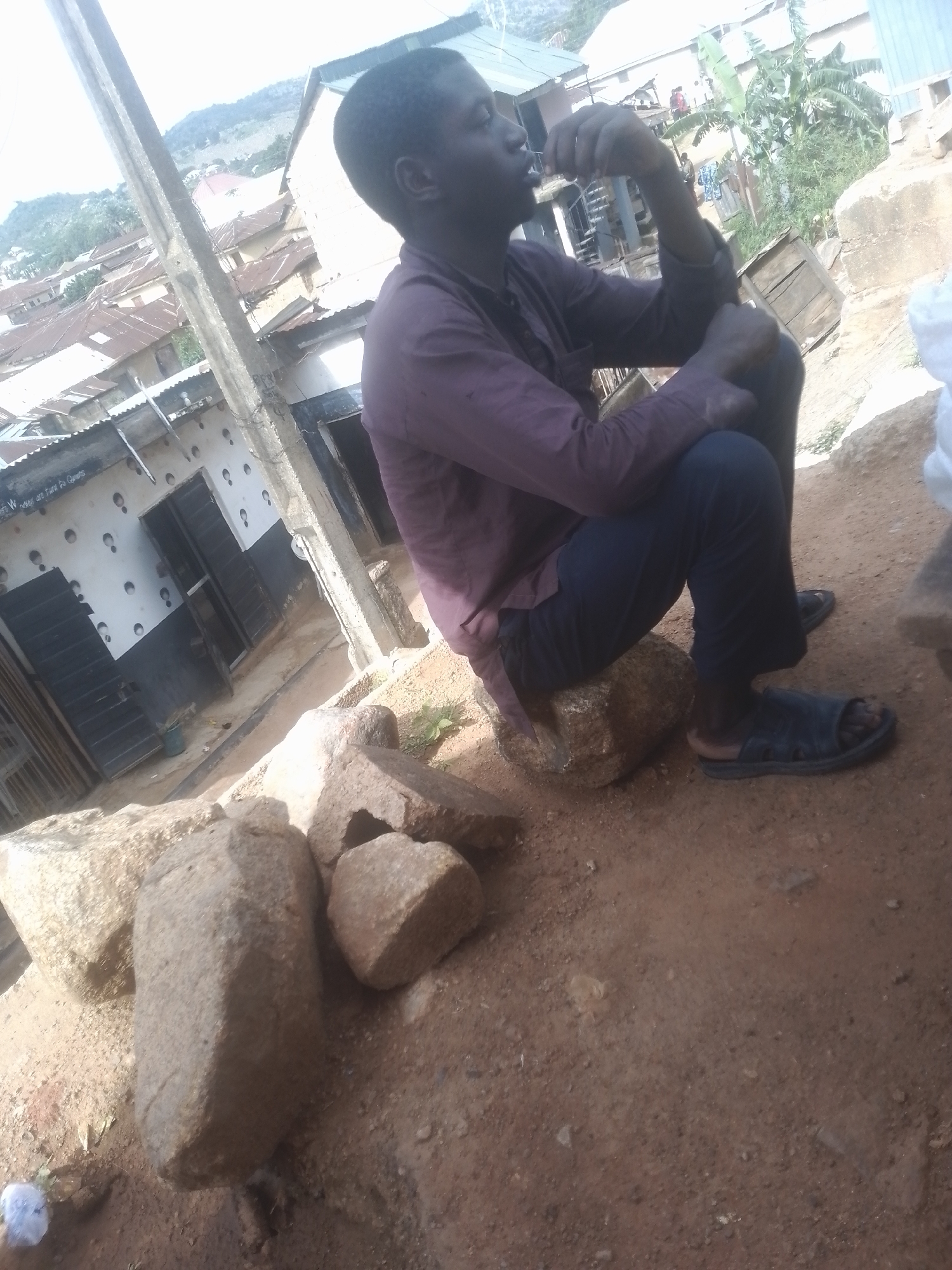 The image shows a young man sitting on rocks in an outdoor setting. He is wearing a dark purple, long-sleeved shirt and dark pants. He has dark skin and short dark hair. He is looking to his left, with his right hand raised to his mouth, as if he is chewing on something or contemplating. He is wearing dark sandals.
The scene appears to be in a somewhat rural or less developed urban area. In the background, there are buildings with corrugated metal roofs and walls made of various materials. Some of the buildings have windows and doors, and there is a large concrete pole in the foreground to the left, which could be a utility pole. There are also several large rocks scattered on the ground around the man. The ground is covered in dirt and pebbles.
The lighting suggests it is daytime, possibly late morning or afternoon, with the sun casting some shadows. The overall atmosphere seems casual and relaxed, with the man appearing to be taking a break or resting. There is a small, blurry white object on the ground in the lower left corner. There is some faint, illegible writing on the wall of one of the buildings in the background. The man's expression is unreadable from this angle.