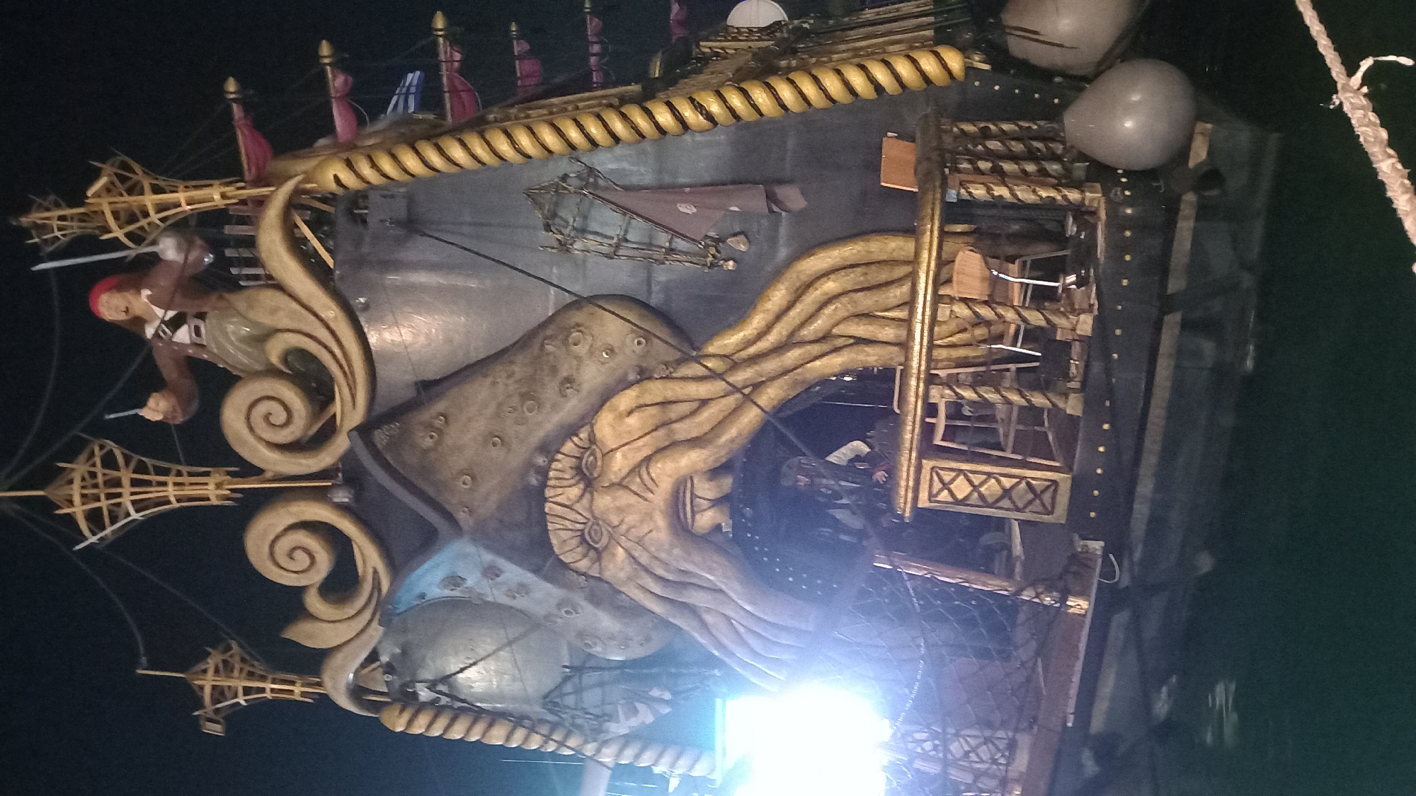 The image shows a large, ornate pirate ship prop, likely from a themed attraction or event, situated on the water at night. The ship is elaborately decorated with a prominent Kraken-like face with tentacles forming an archway in the center of the stern. Above the archway, a male figure dressed as a pirate, wearing a red bandana and brown jacket, stands with two swords raised. The ship features swirling golden decorative elements, a thick rope detailing along the side, and various wooden structures, including railings and masts. Red and blue sails are visible in the background. In the lower part of the image, the dark hull of the ship is visible, partially submerged in dark water. The scene appears to be illuminated by artificial lights, with some areas, particularly on the left side, exhibiting bright lens flare. The ship is located at the Kos Marina in Kos, Greece, a common location for boat moorings and potentially for such attractions.